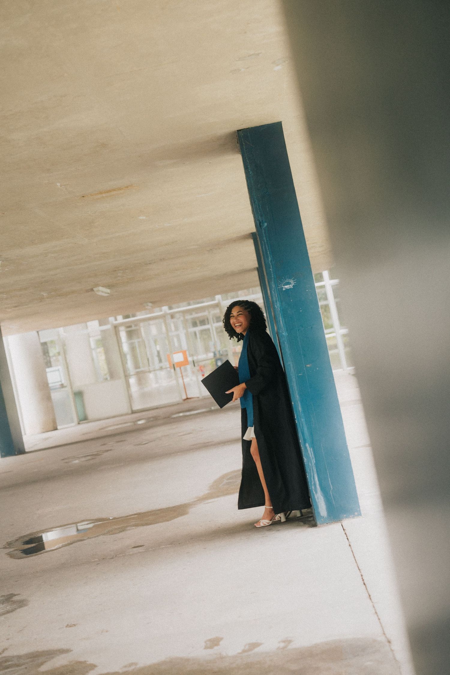 A figure dressed in black stands near a blue pillar in an empty concrete parking structure with natural lighting.