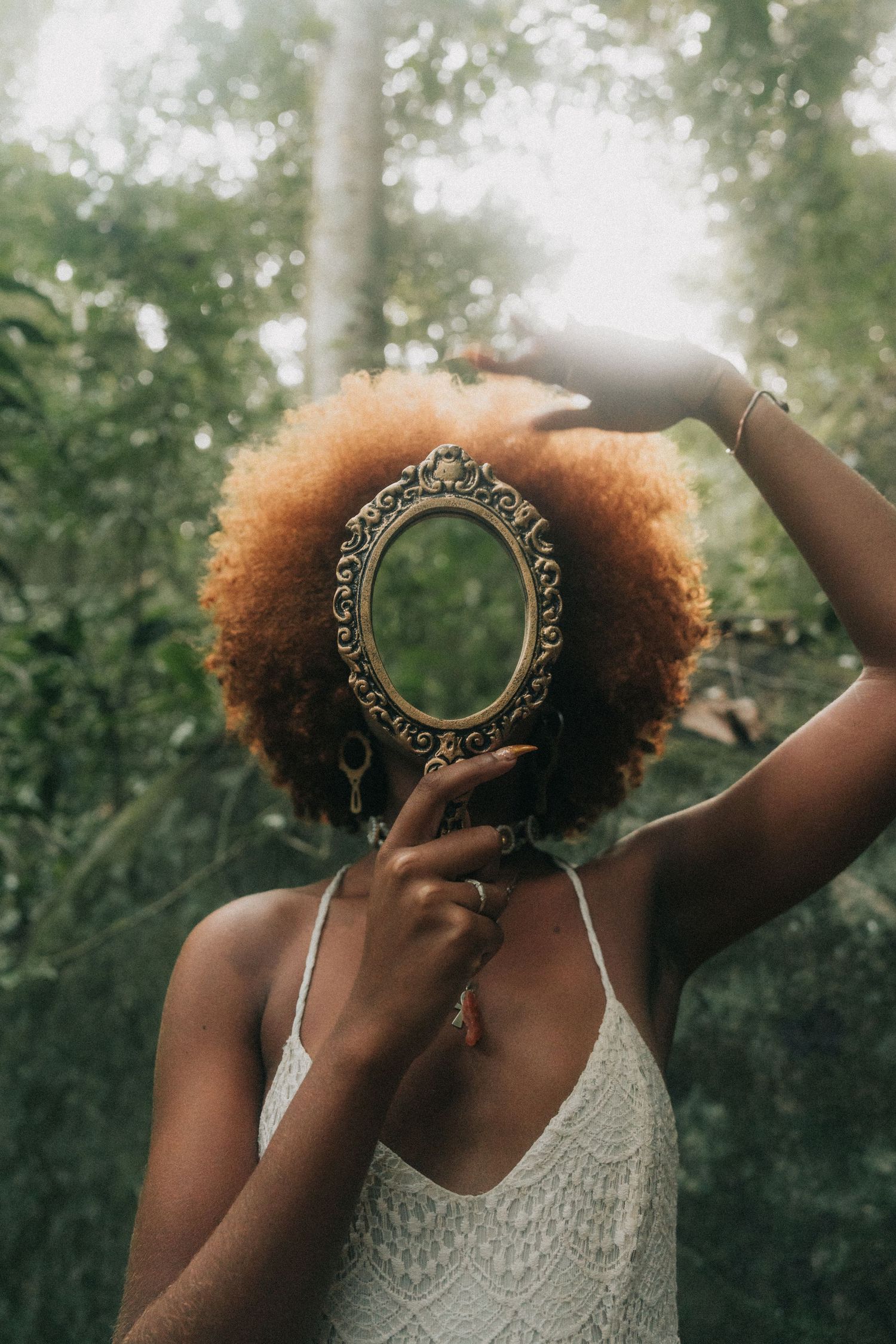 A person in a white lace dress holds up an ornate vintage mirror in a dreamy outdoor setting.