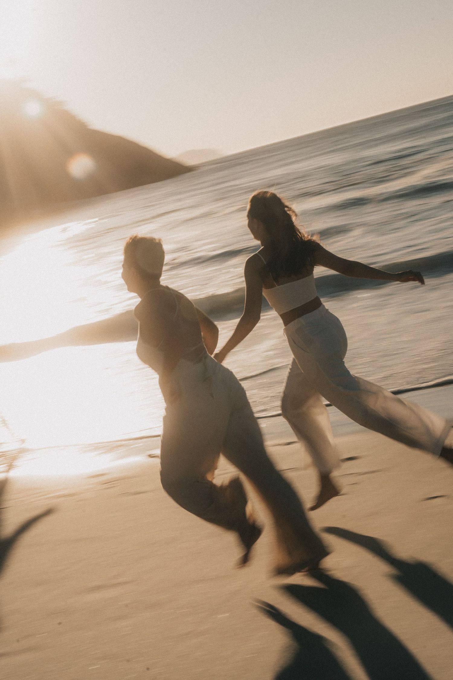 Two silhouettes run along a sunlit beach shore as waves crash nearby, creating a dreamy sepia-toned scene.