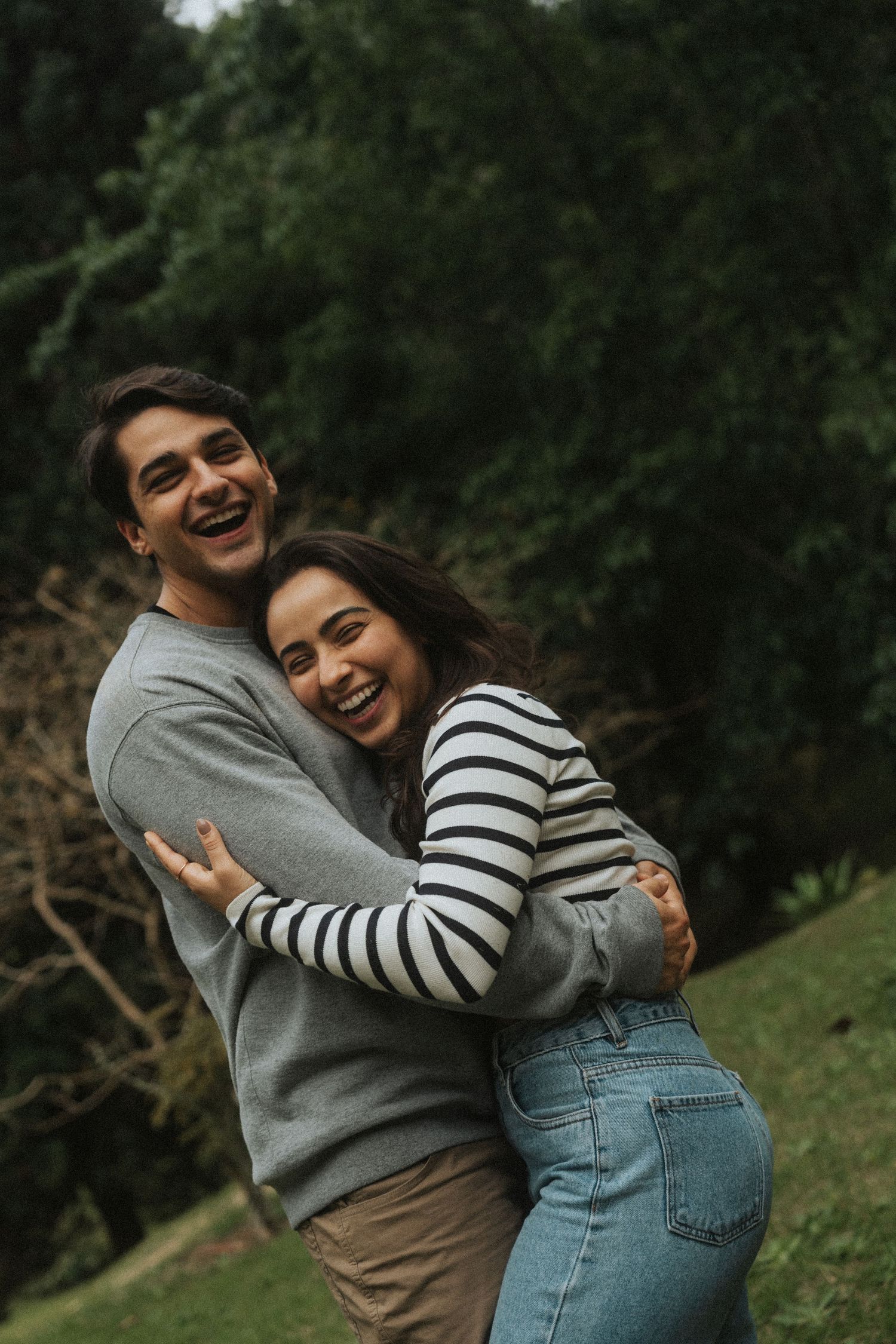 A couple in casual attire laughing and embracing outdoors in a natural setting with trees in the background.