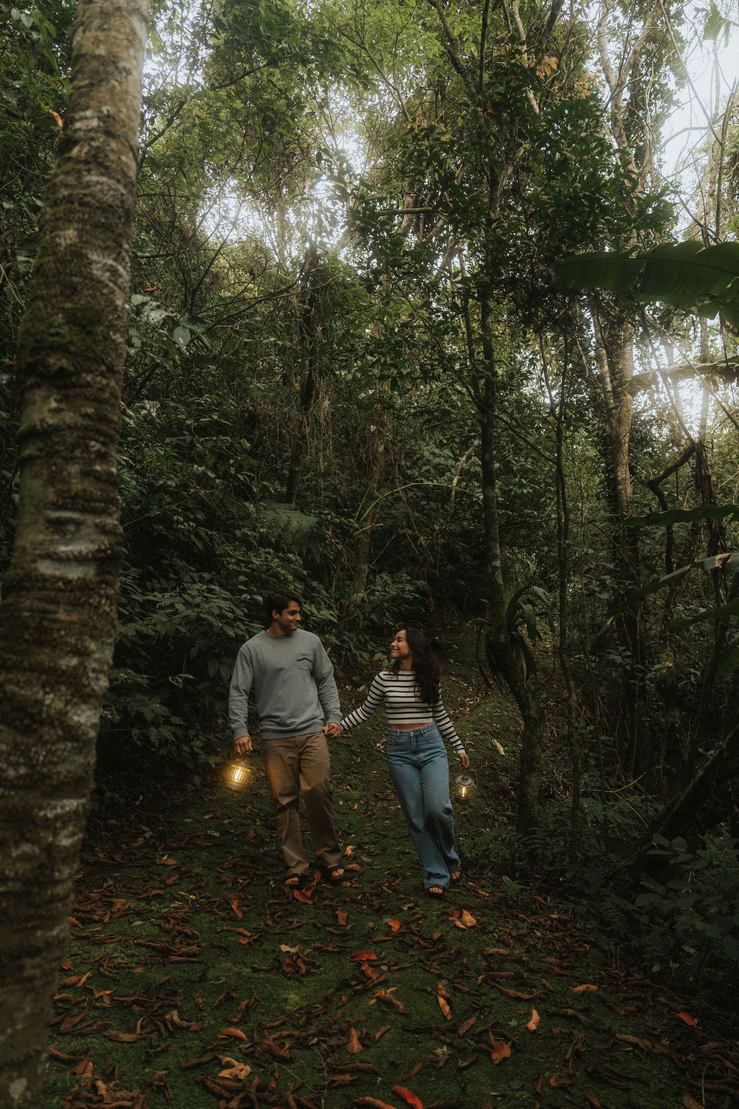 Two people walk along a wooded path with scattered orange pumpkins during autumn season.