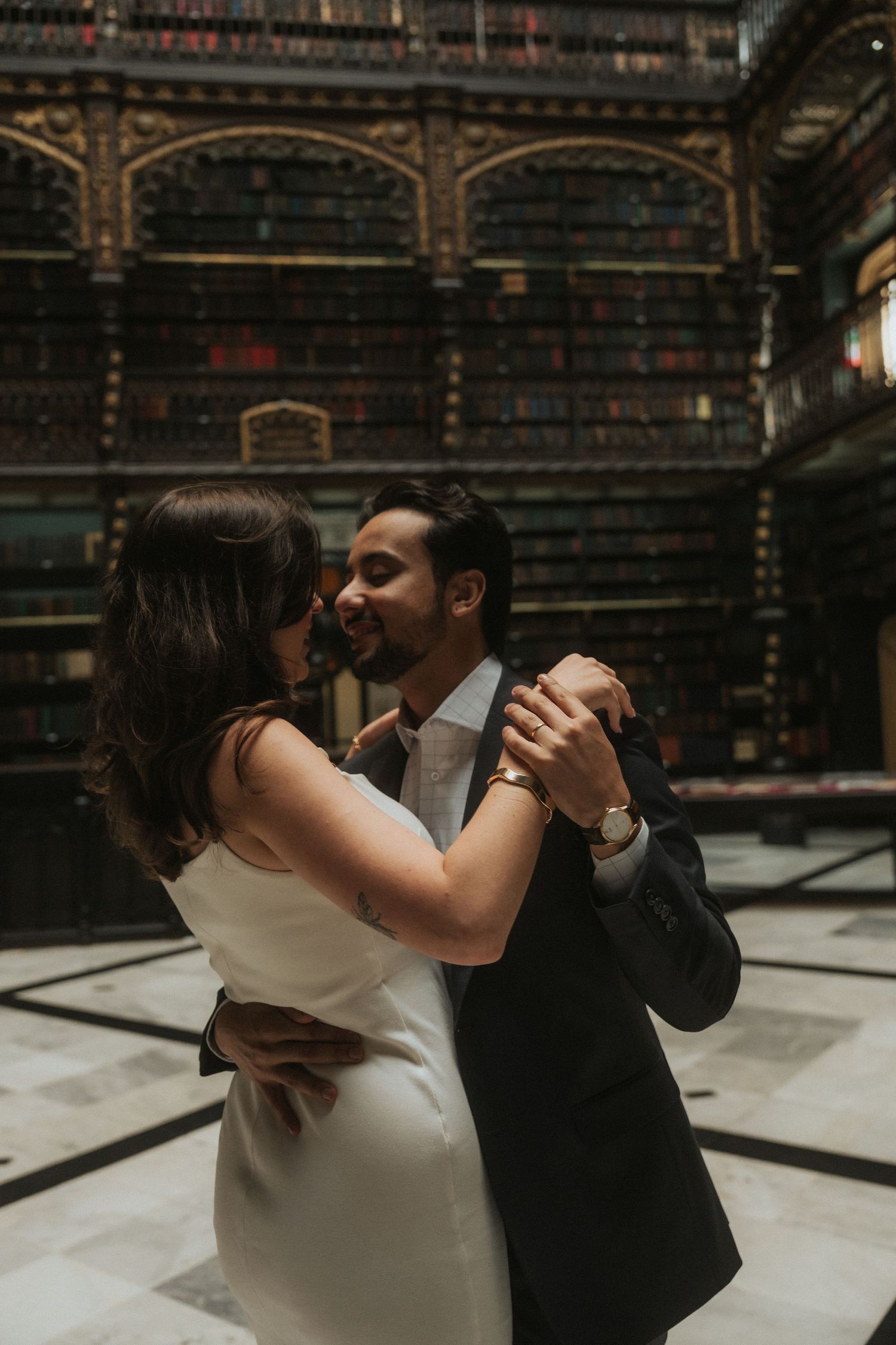 A couple shares an intimate dance in an elegant library with ornate arched bookshelves and classic architecture.