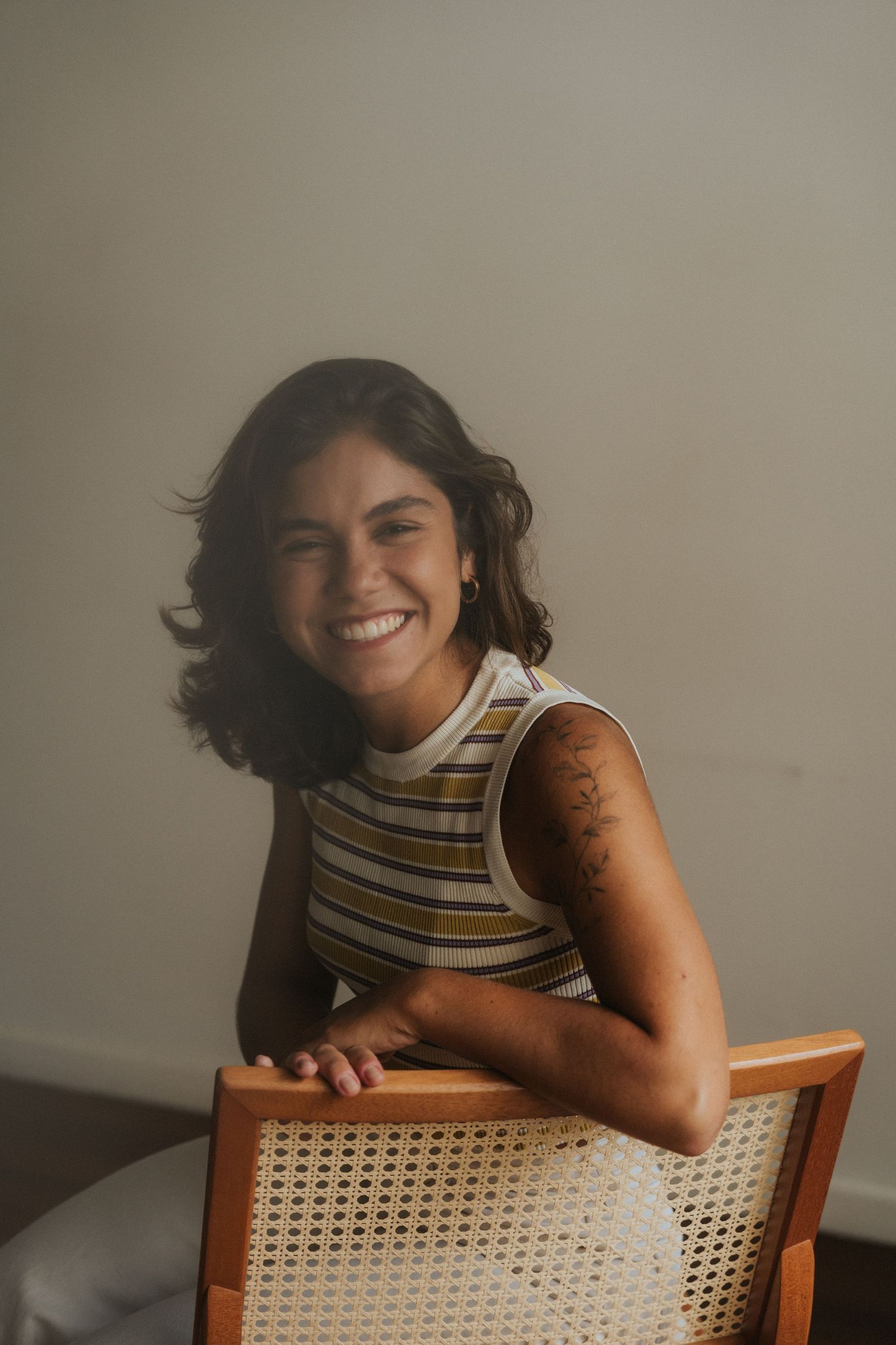 A smiling person in a striped tank top sits casually in a cane back chair against a neutral background.