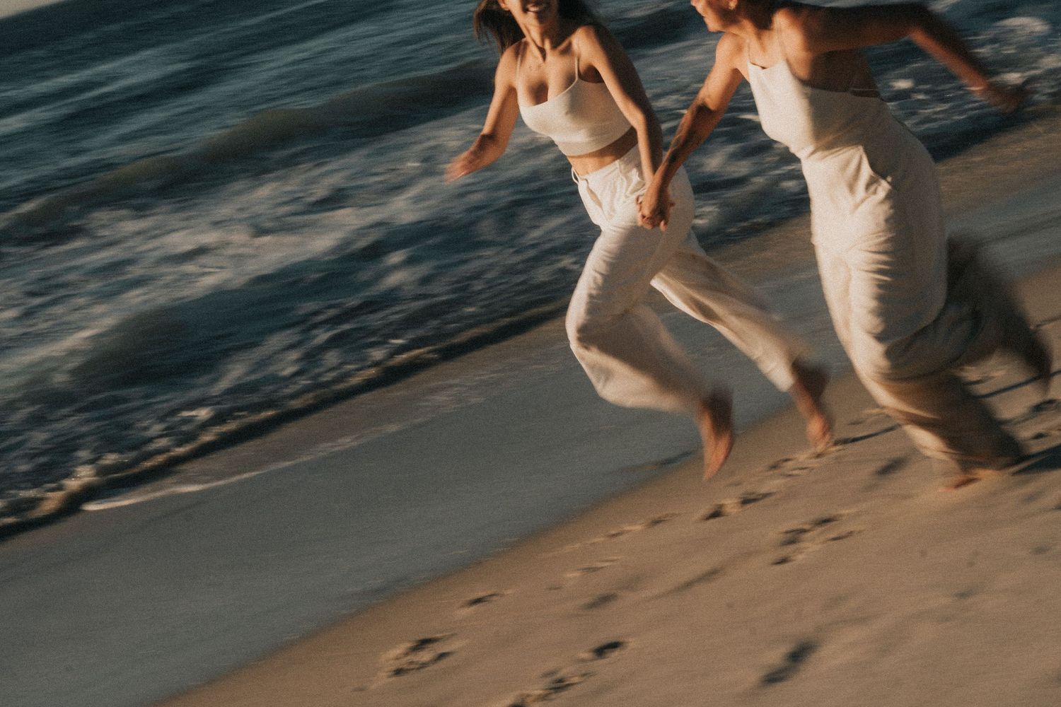 A couple walks along a sandy beach at sunset holding hands, leaving footprints in the wet sand.