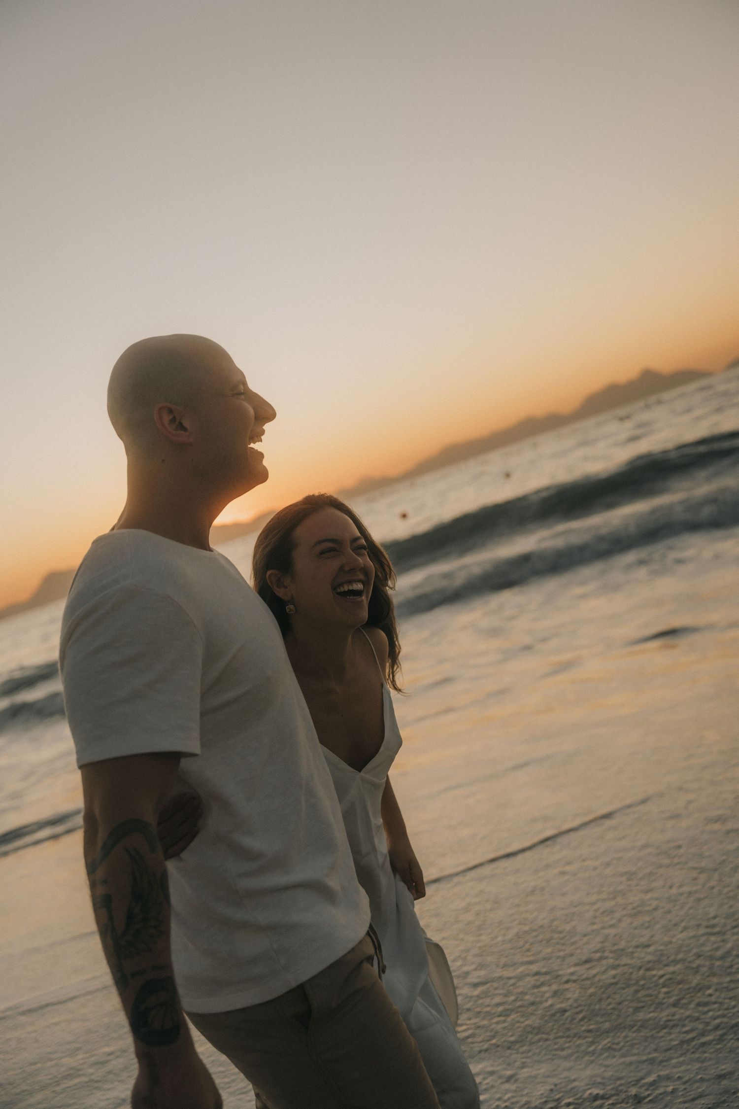A couple embraces on the beach during a romantic sunset with ocean waves in the background.