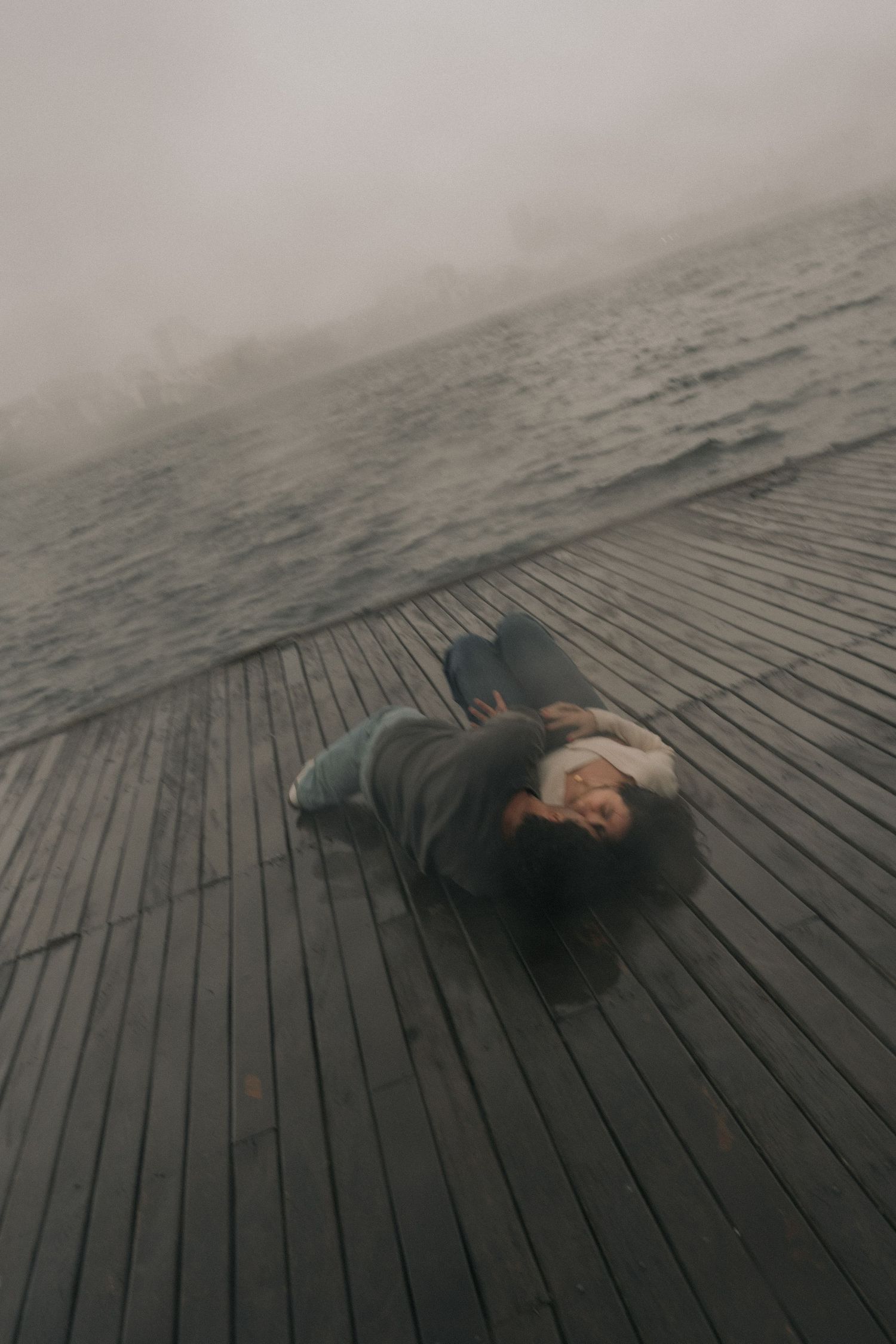 A person lies curled up on wooden boardwalk planks near foggy water on a misty gray day.