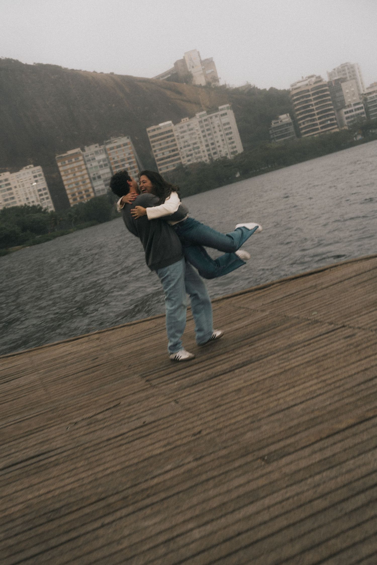Two people embrace and dance romantically on a wooden dock by a lake with apartment buildings visible in the background.