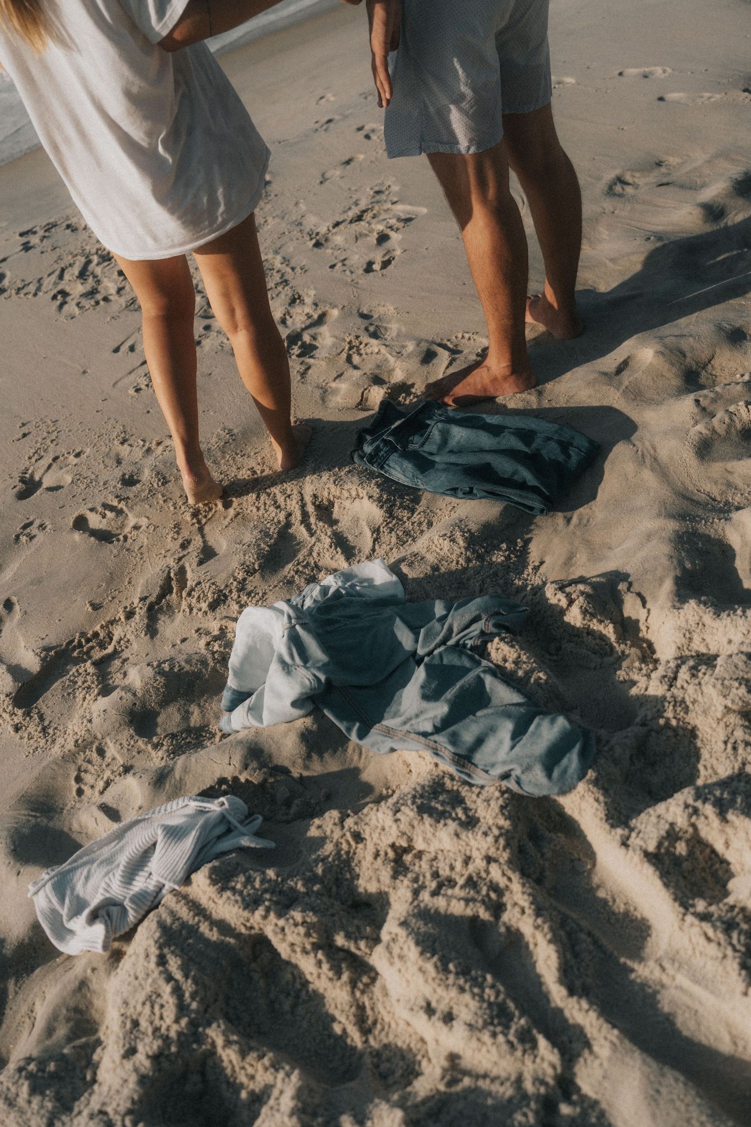 Bare legs standing on beach sand next to scattered blue denim clothing items in warm sunlight.