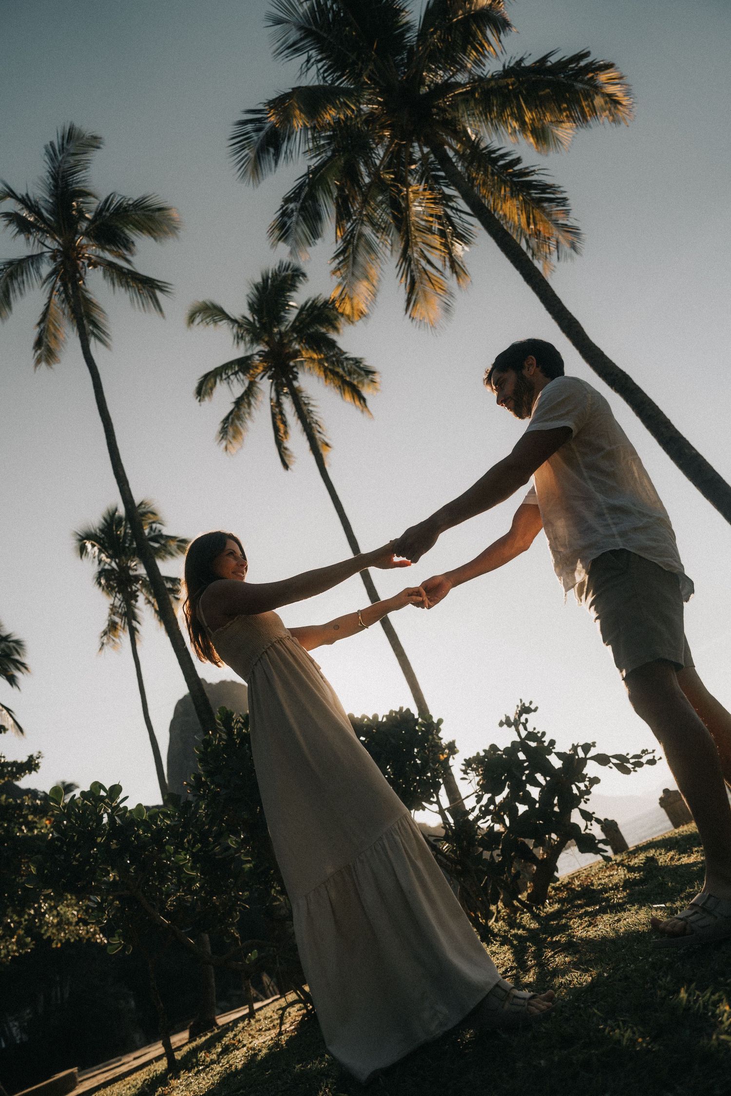 Silhouetted figures hold hands against sunset sky with tall palm trees swaying in tropical paradise.