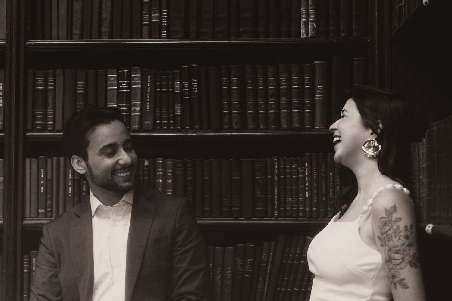 A black and white photo shows a couple in formal wedding attire posing in a library setting.