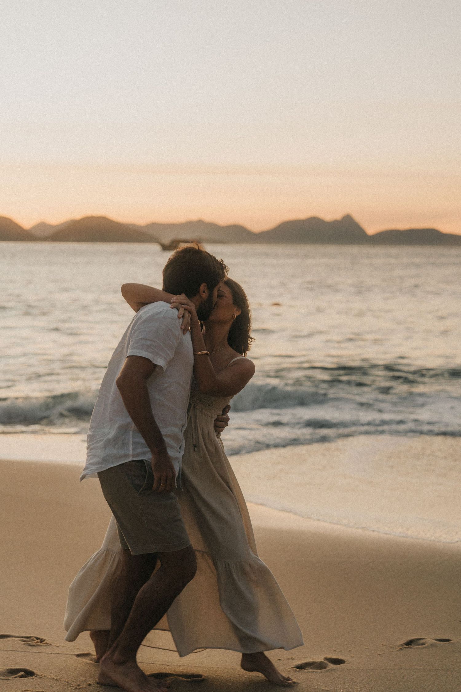 Romantic couple embracing on sandy beach at sunset with mountains and ocean in background.