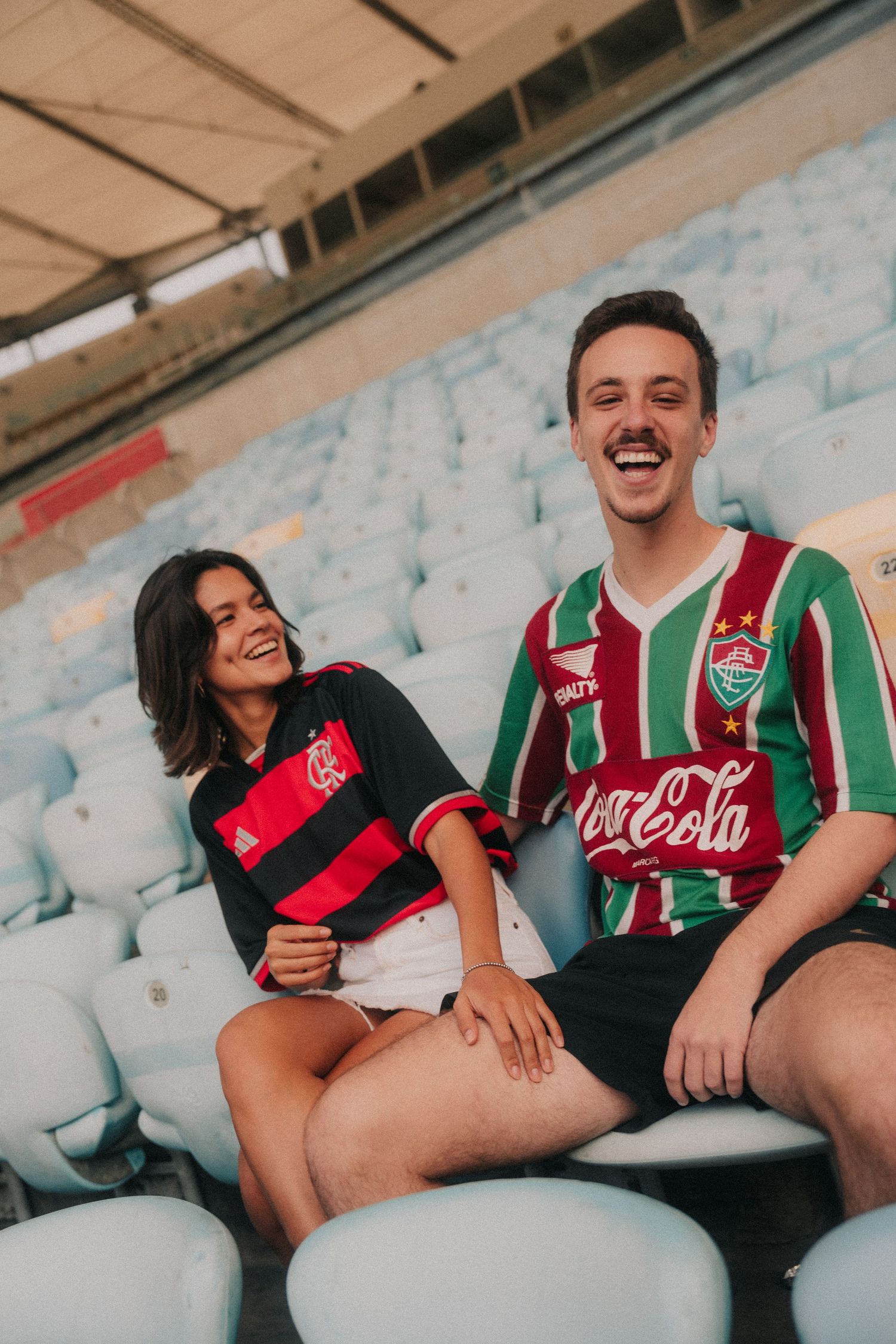 Two soccer fans wearing Flamengo and Fluminense jerseys sitting together sharing a happy moment.