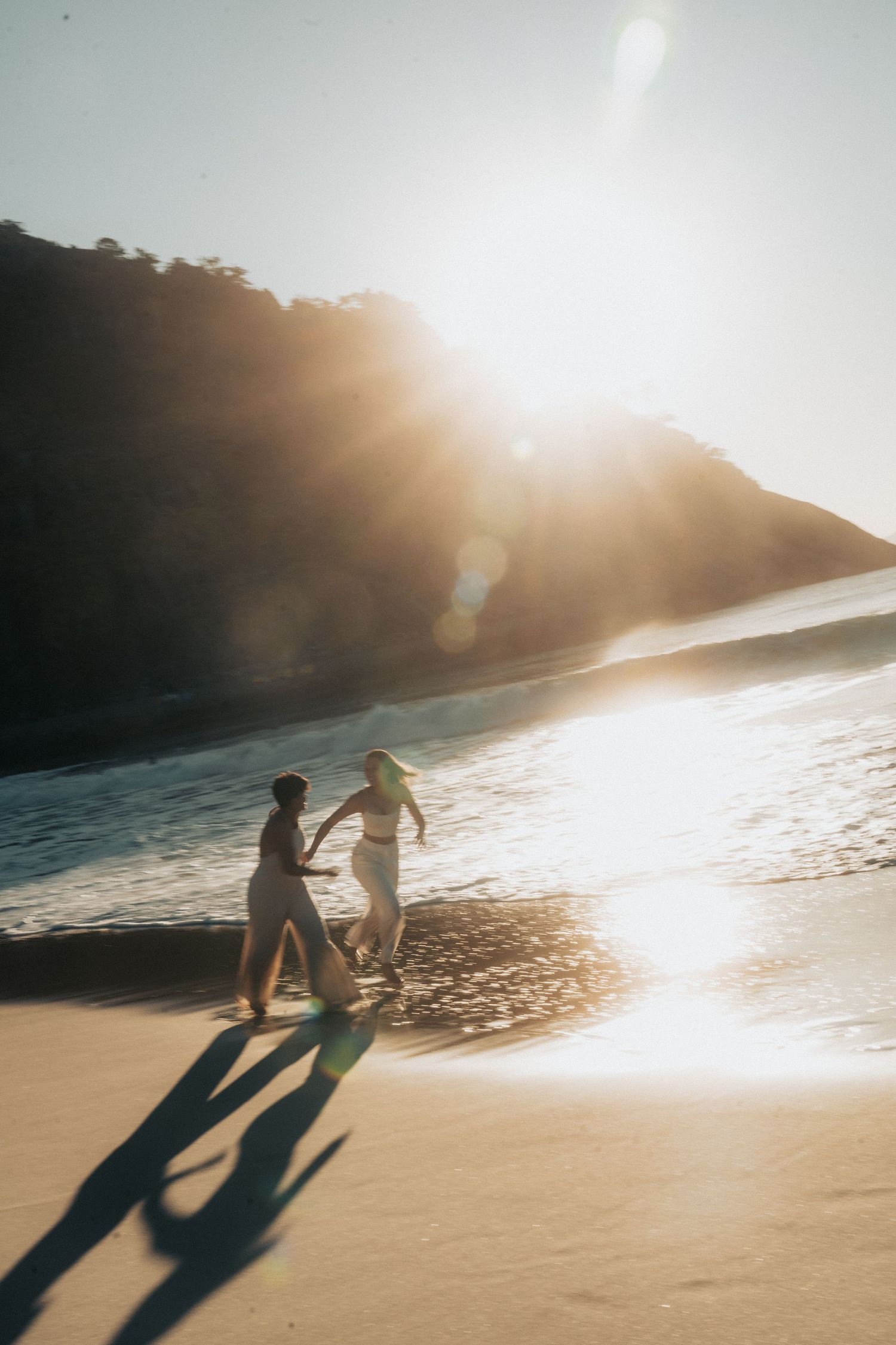 Two silhouettes dance playfully on a sunlit beach during golden hour, casting long shadows across the pristine sand.