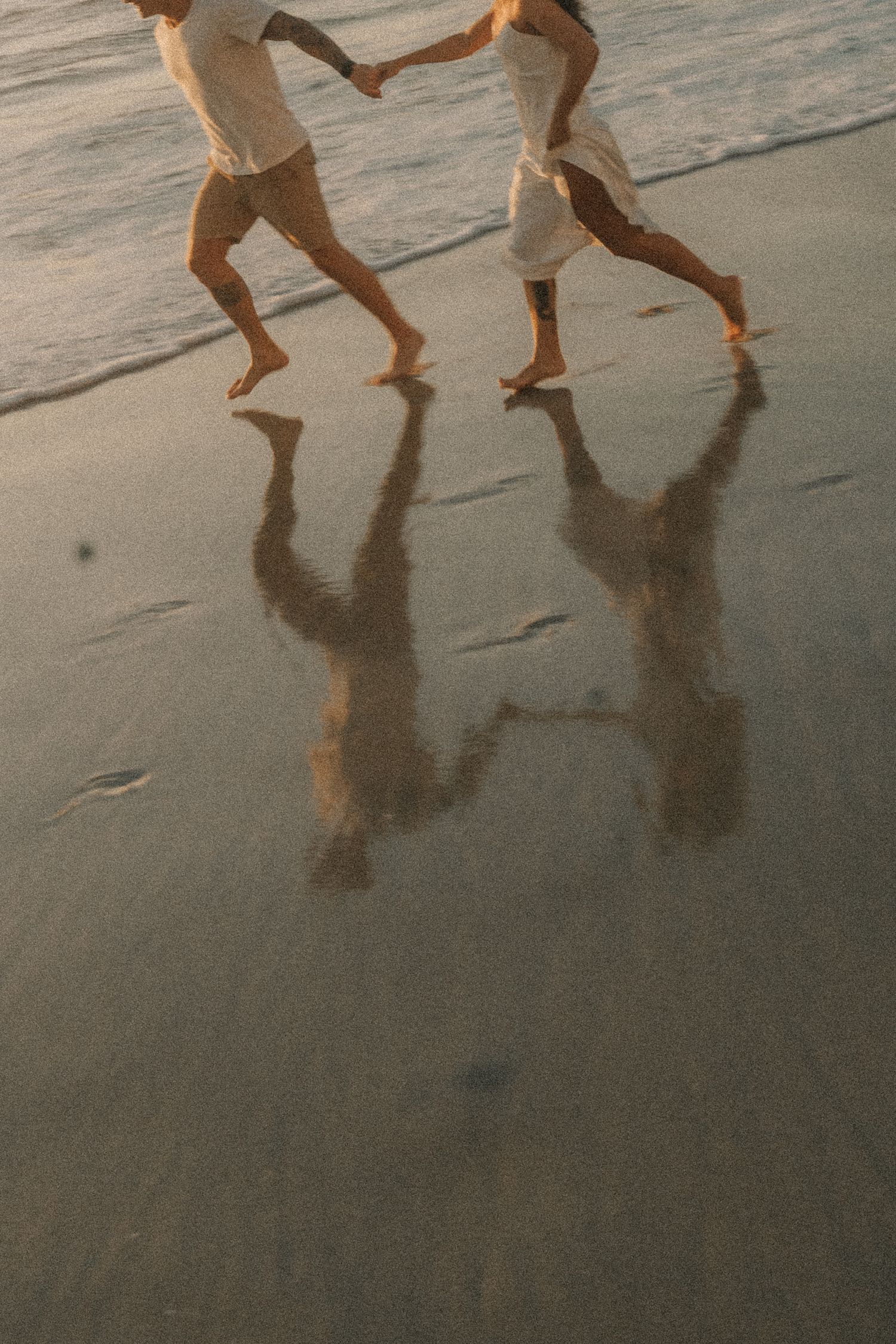 Silhouettes of bare feet and legs running on wet sand at dusk, creating mirror-like reflections on the beach.