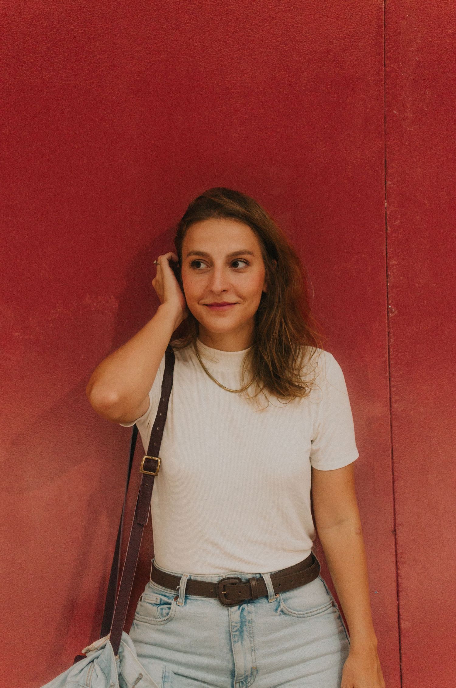 Person in a white t-shirt and light denim jeans poses against a red wall background.