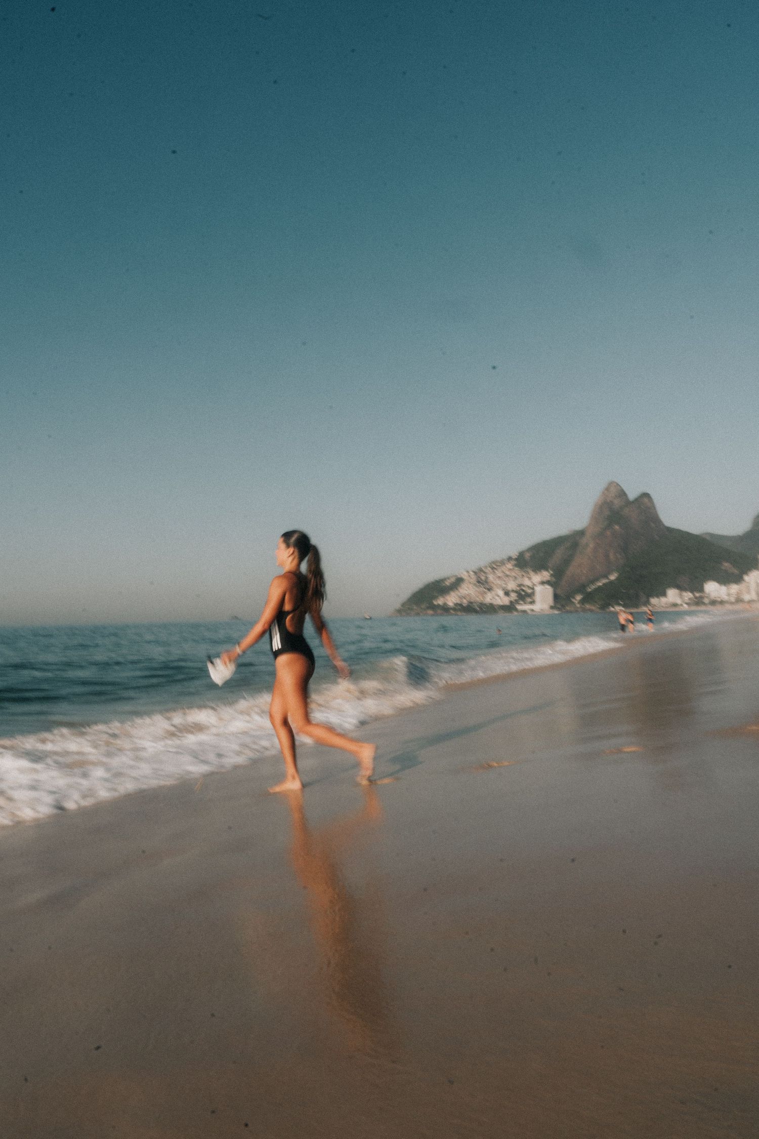 Woman playing in the waves at a scenic beach with mountains and coastal buildings in the background.