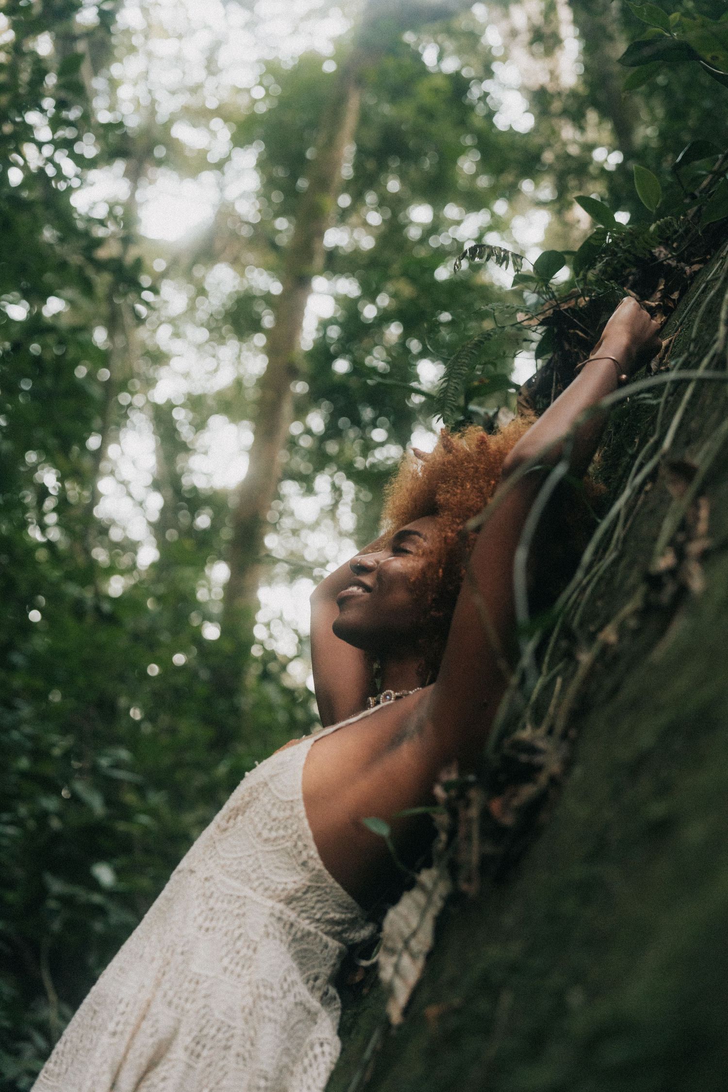 A joyful person in a white lace dress laughs while surrounded by lush green foliage in a sunlit forest setting.