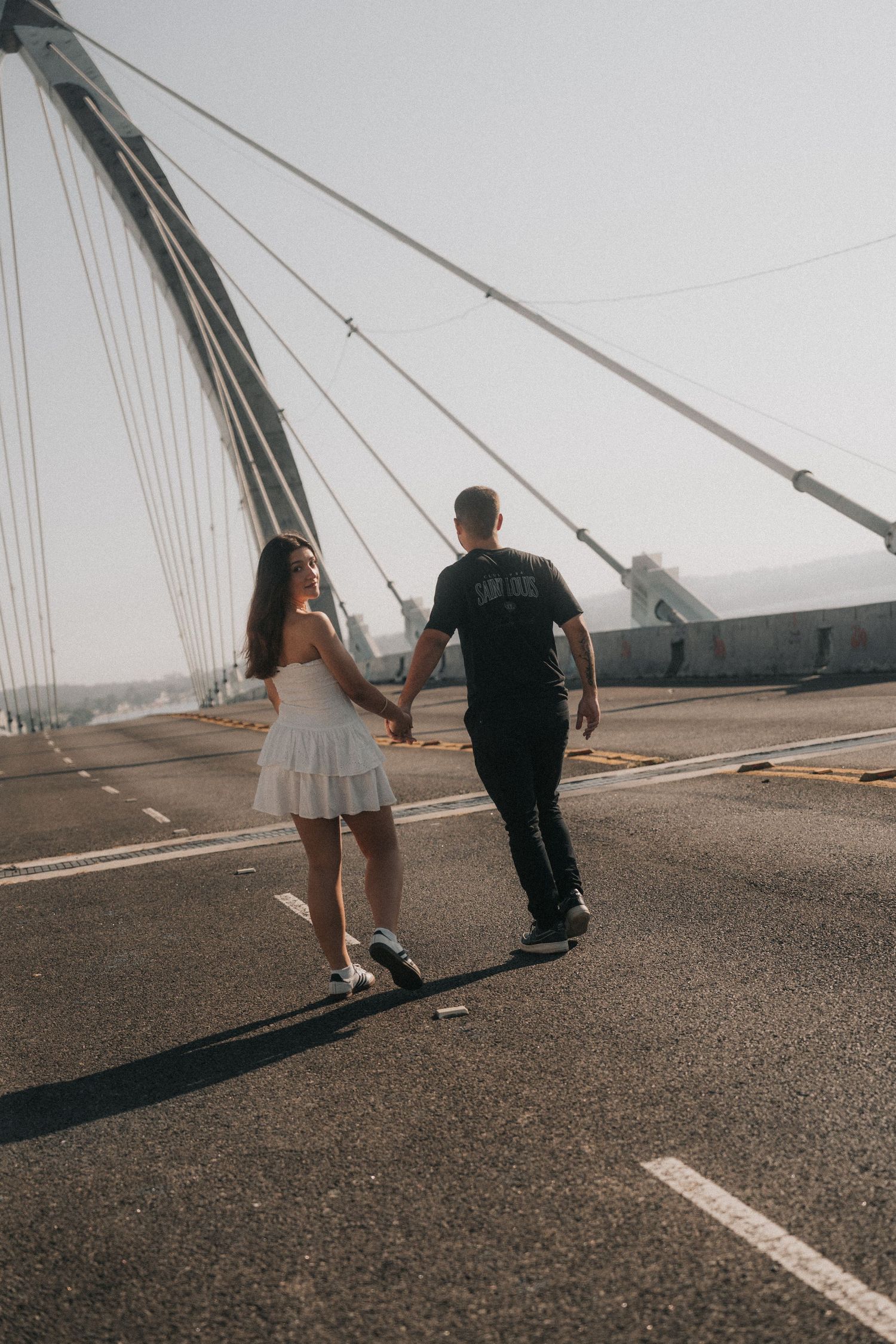 A couple walking away on an asphalt surface near angled cable or pole structures creating dramatic shadows.