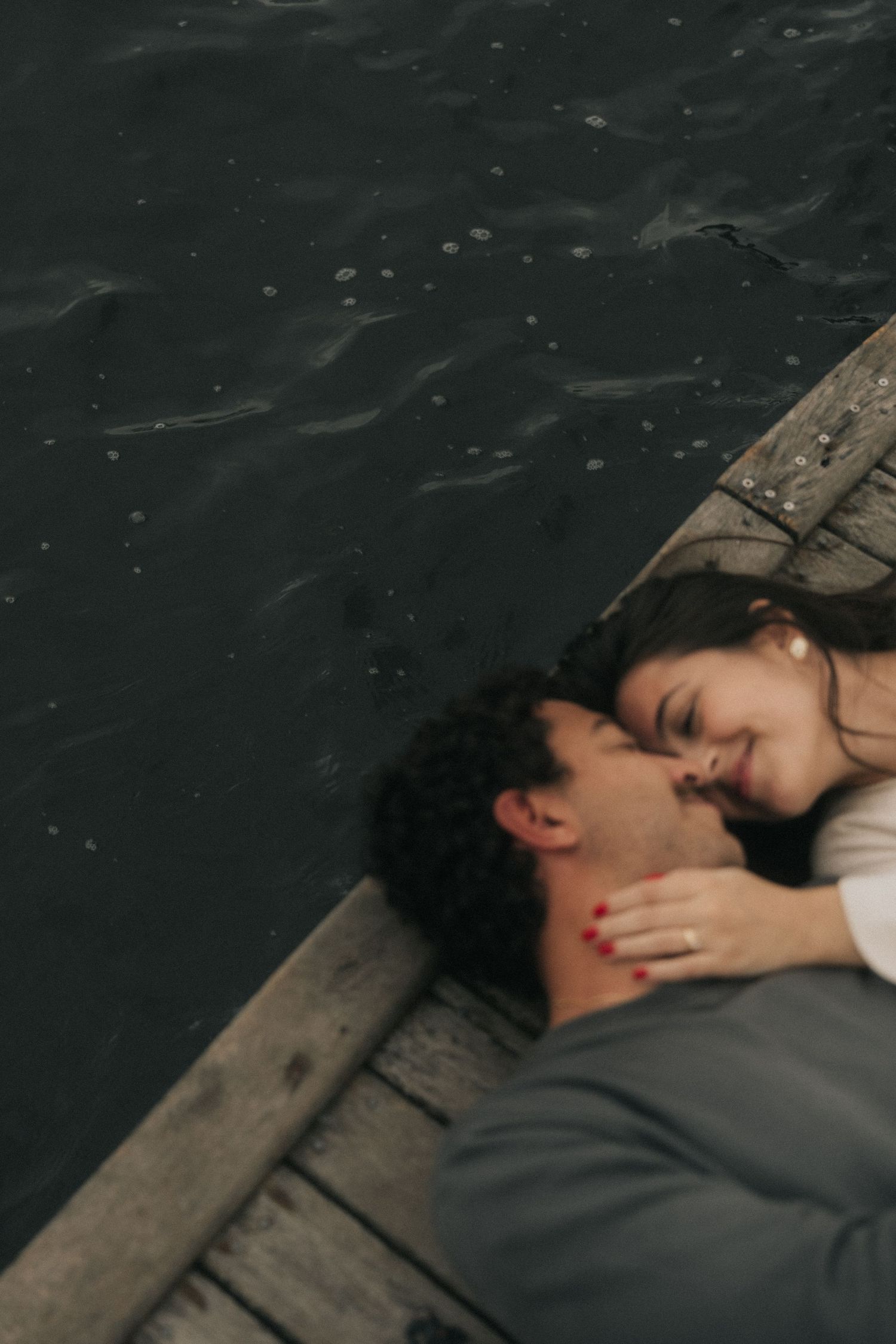 A couple shares an intimate moment lying together on a wooden dock above dark waters.