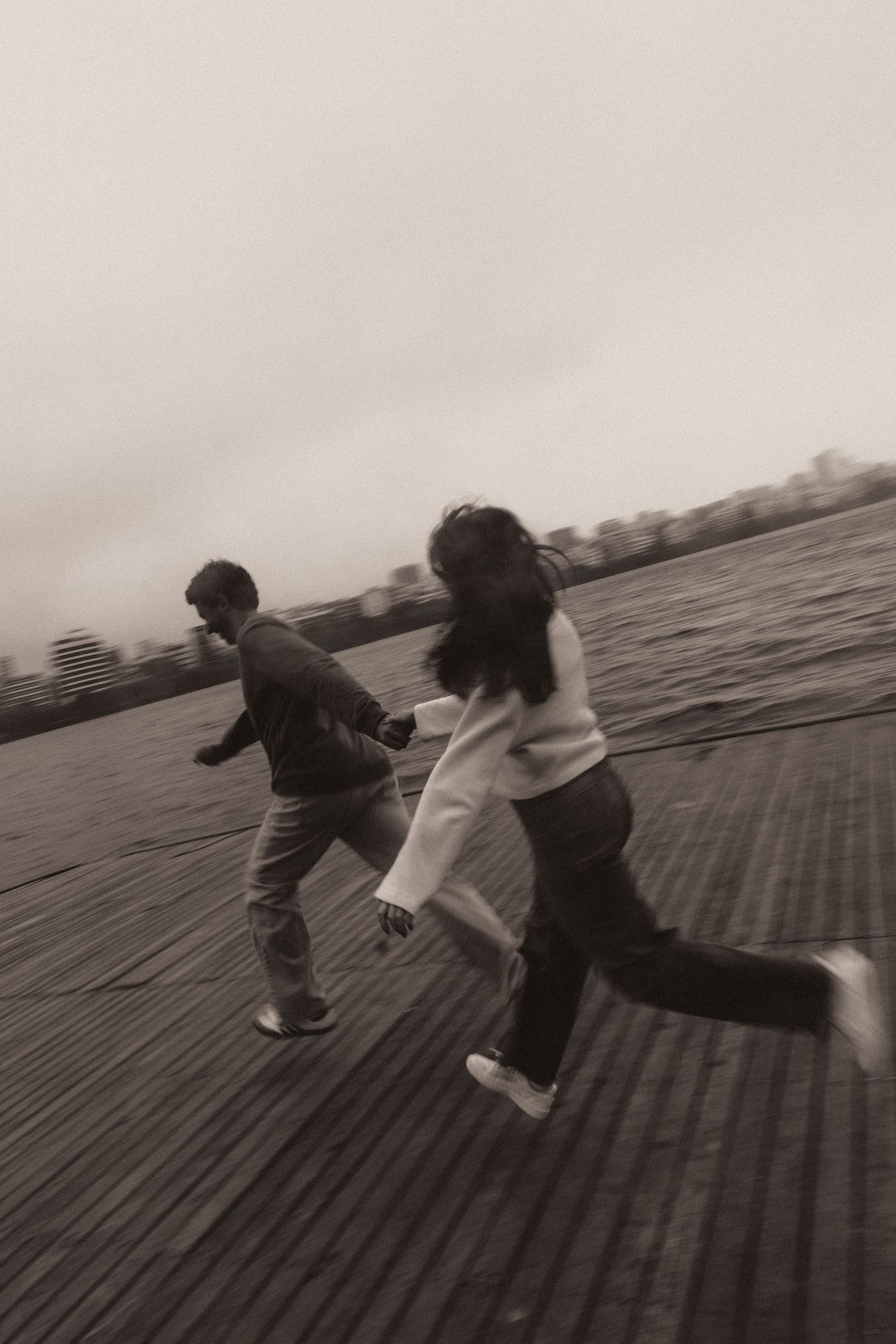 Two people playfully run together across a wooden boardwalk in a black and white motion sequence.