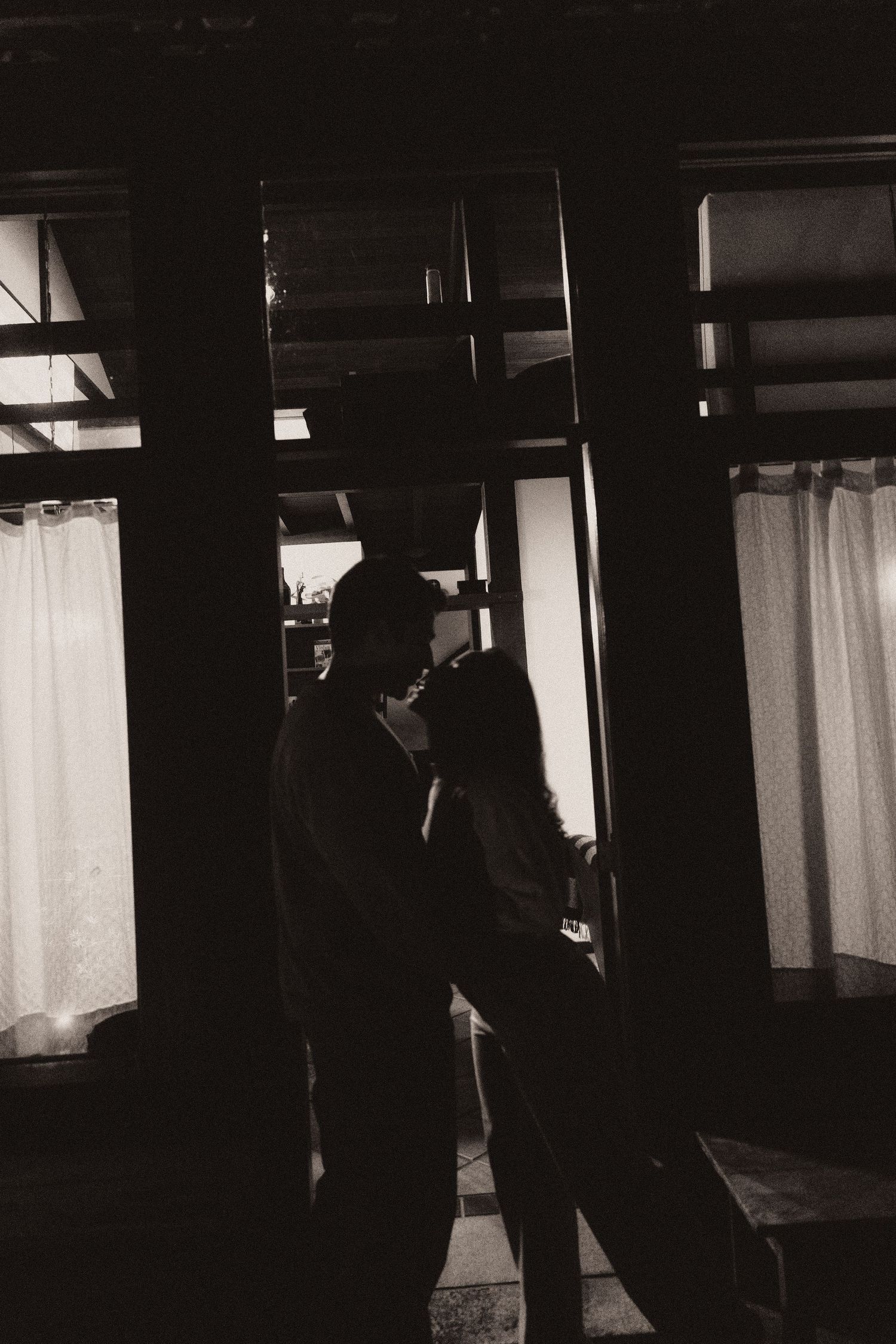 A moody black and white photograph showing silhouettes standing in a dark interior space with curtained windows.