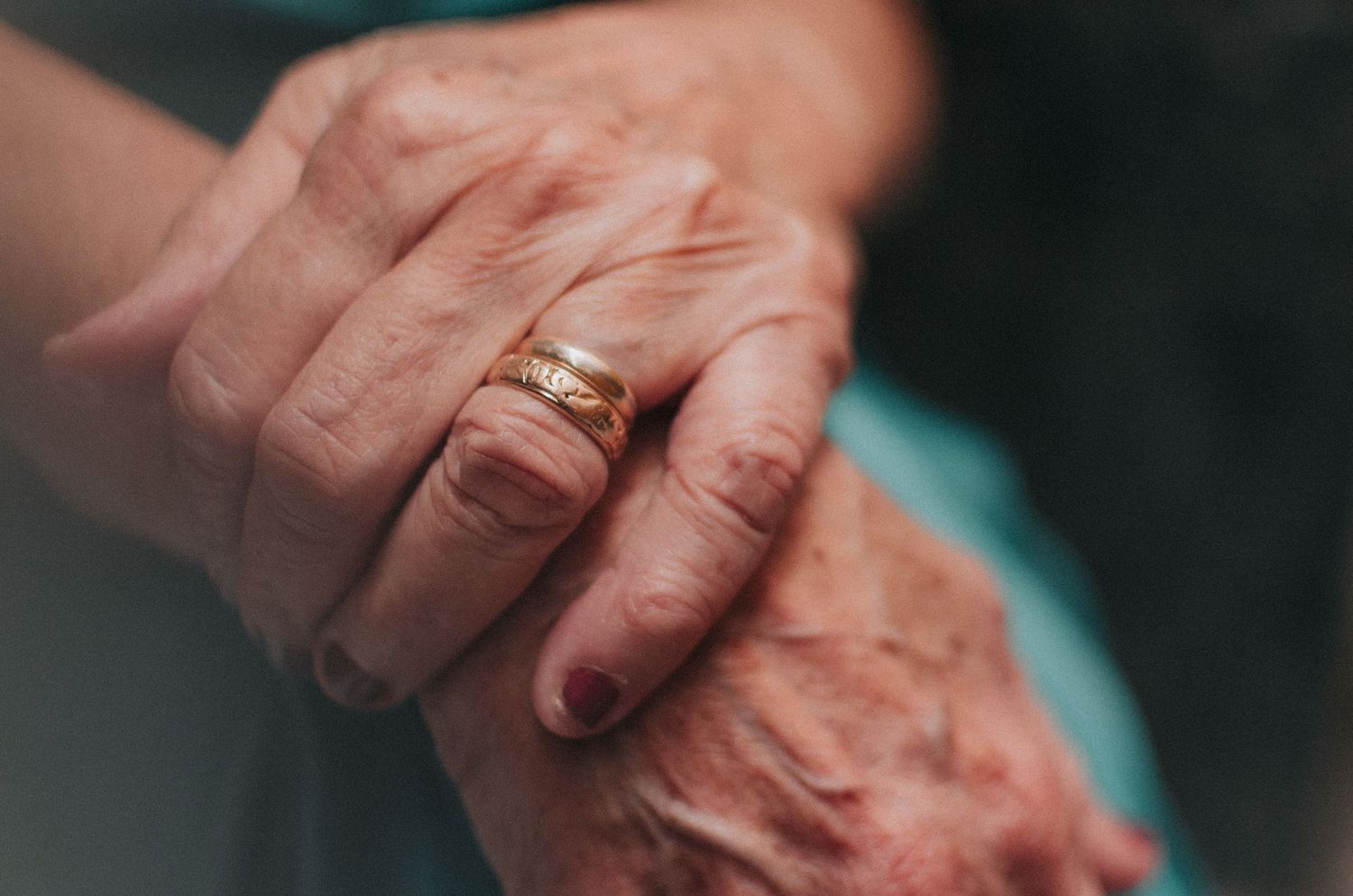 A pair of elderly hands wearing wedding rings holds each other in a tender close-up moment.