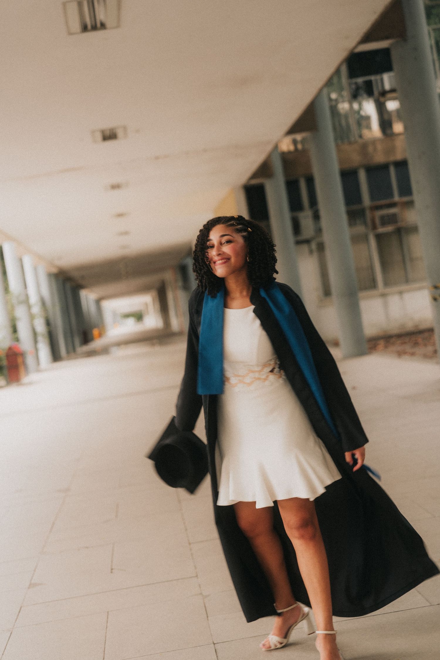 A person in a long navy coat and white dress poses in different angles along a white corridor.