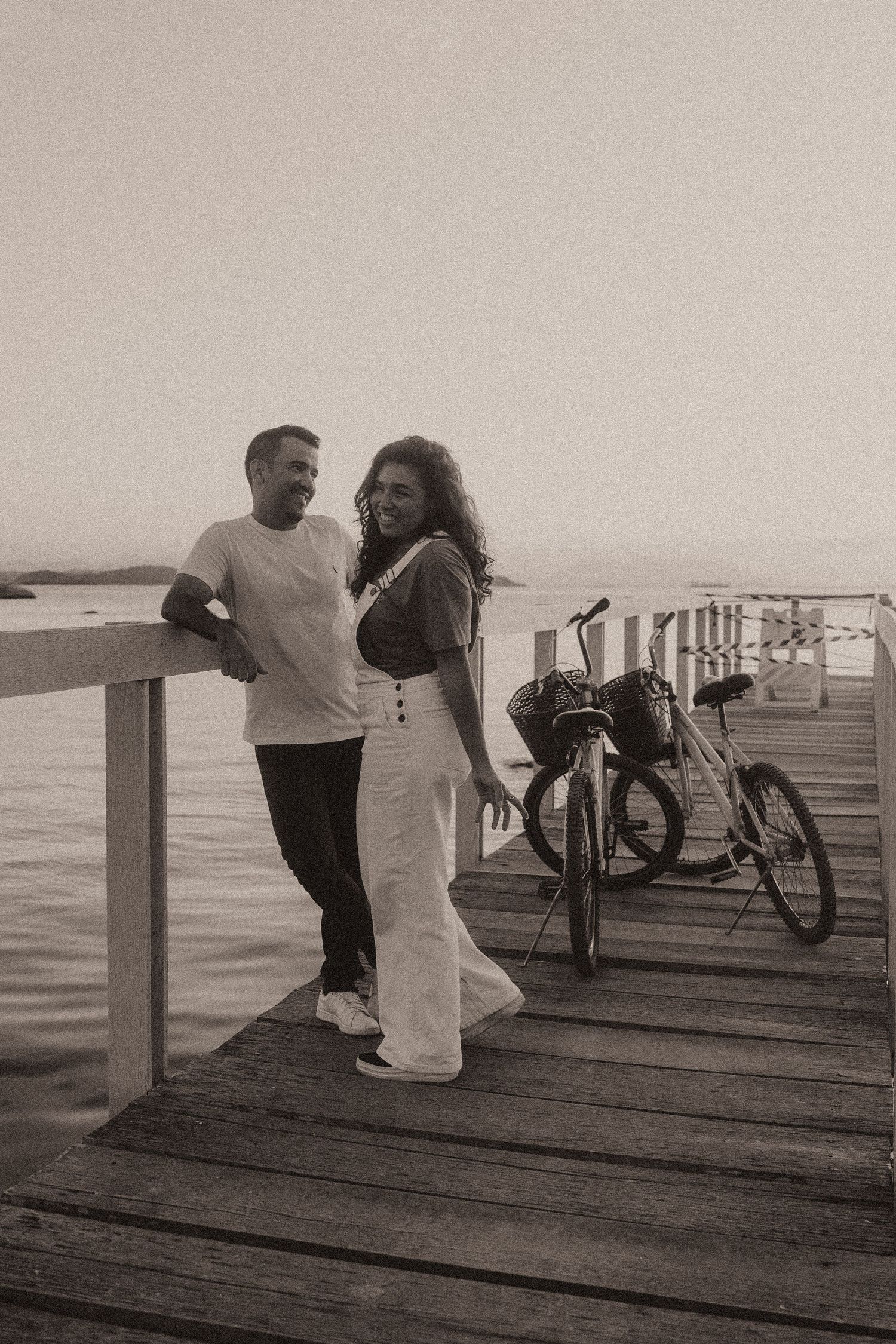A couple sits together on a wooden pier next to their bicycle, enjoying a peaceful oceanfront view in black and white.