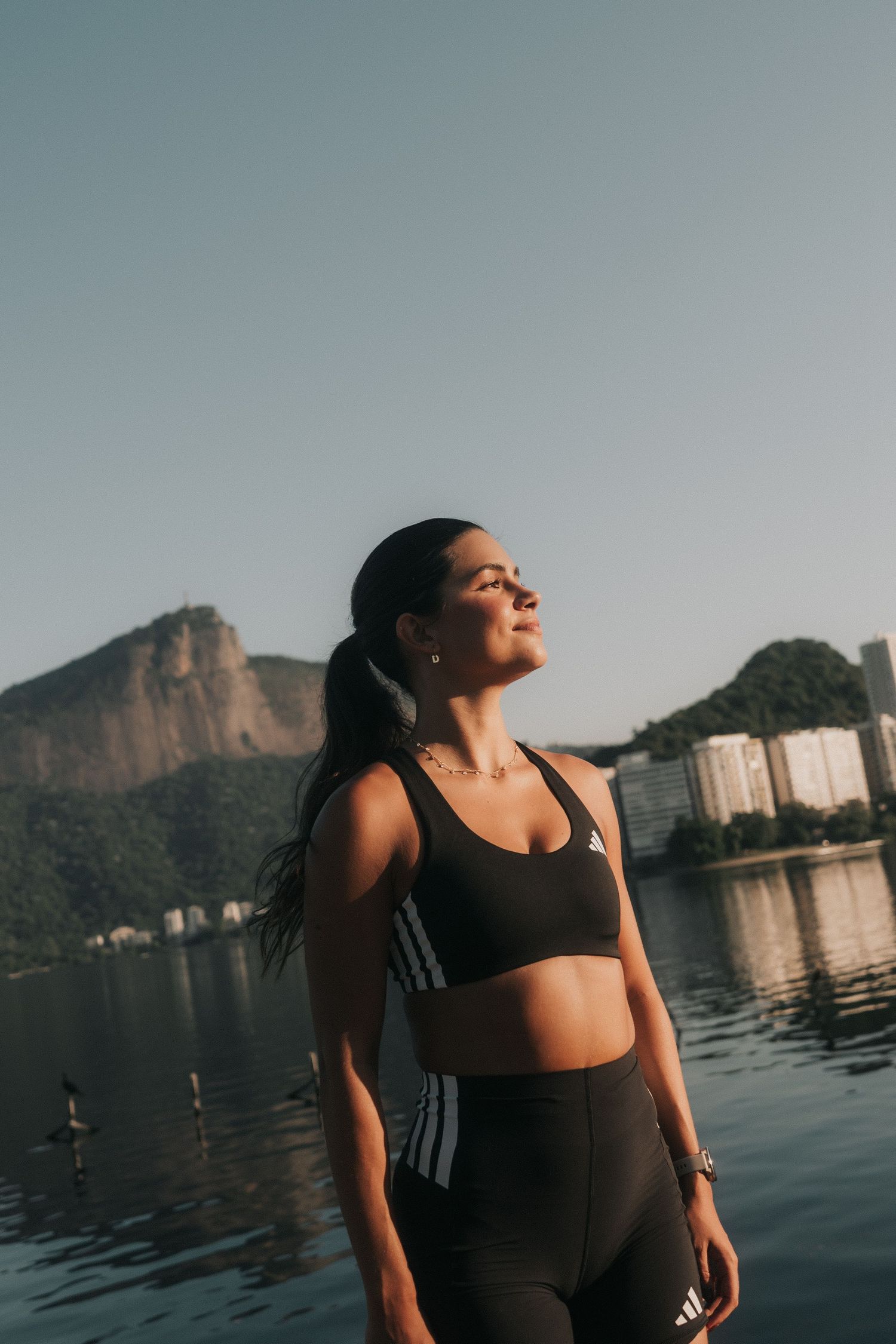 Athletic woman in black sportswear poses against scenic waterfront backdrop with mountain and cityscape at sunset.