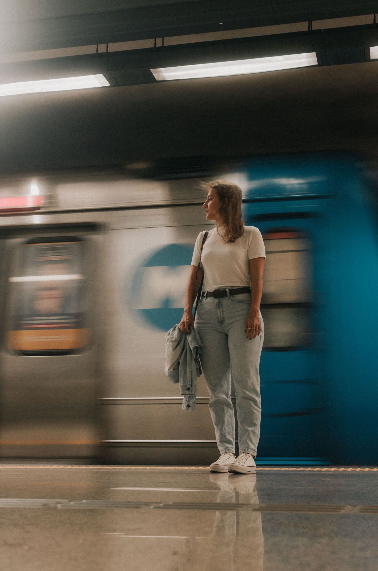 Person in casual wear stands on subway platform as blurred train motion creates dynamic urban transit scene.