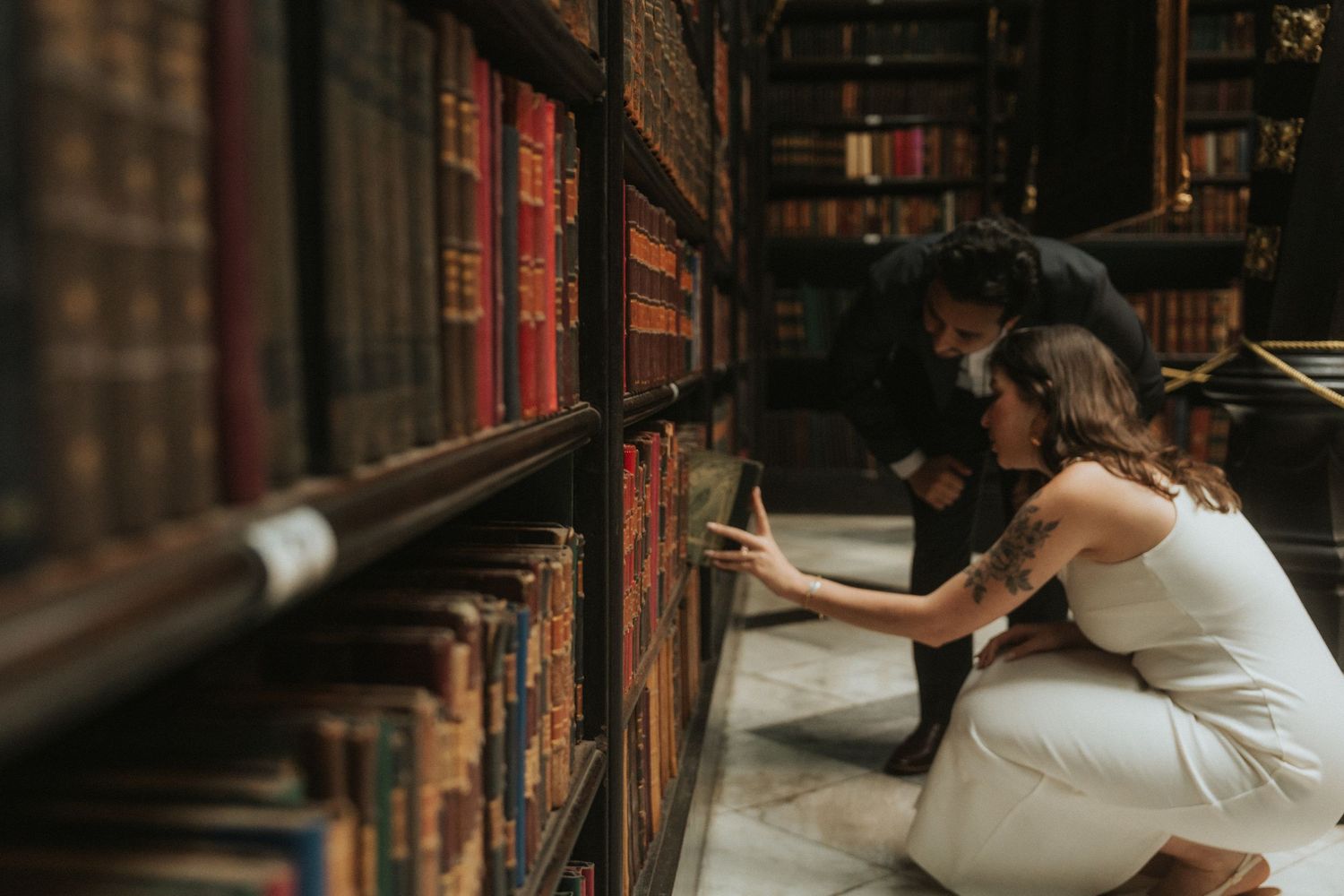 Two people in white outfits browse old books on wooden library shelves in a dimly lit vintage library setting.