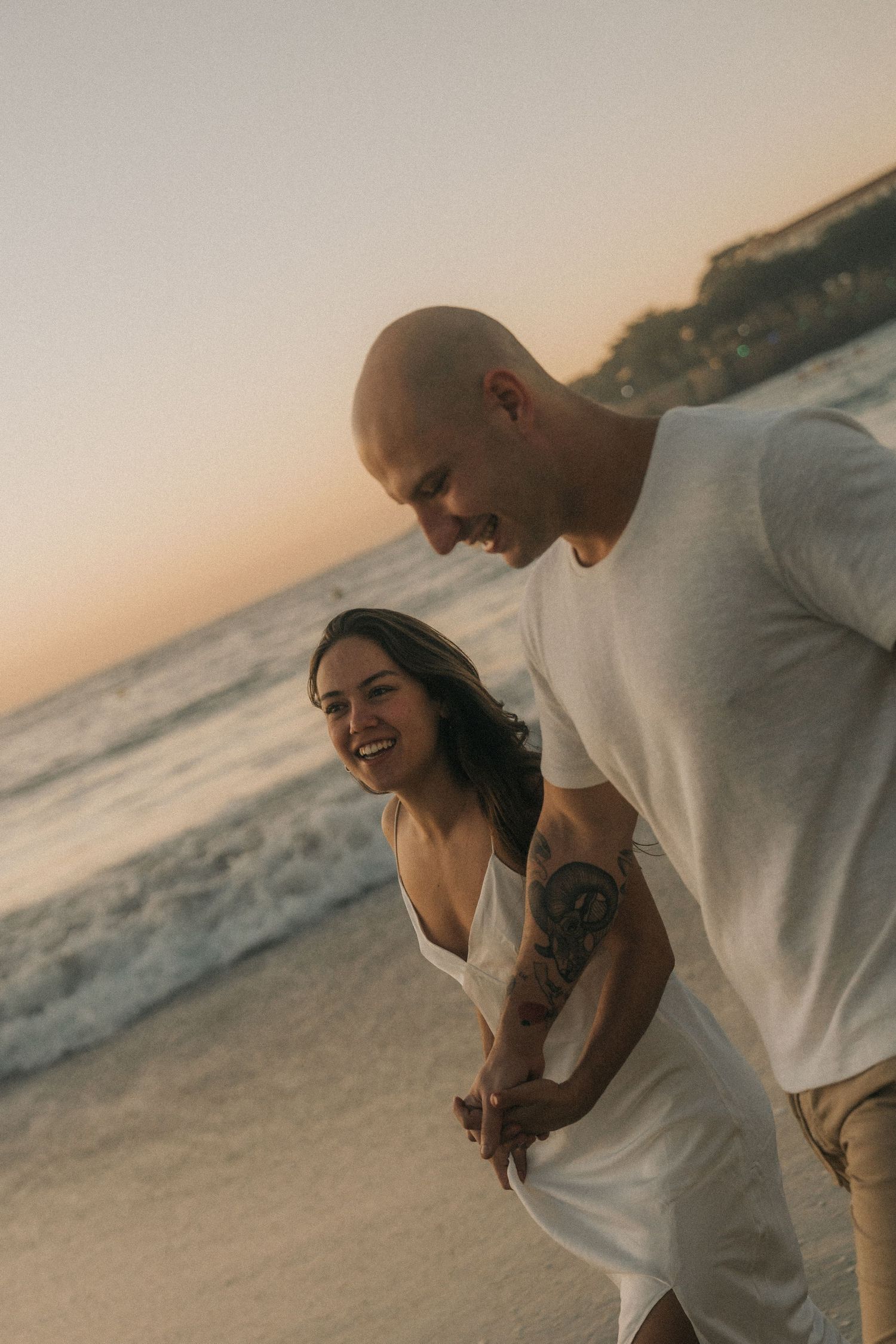 A couple embraces affectionately on a sandy beach during sunset while waves crash in the background.