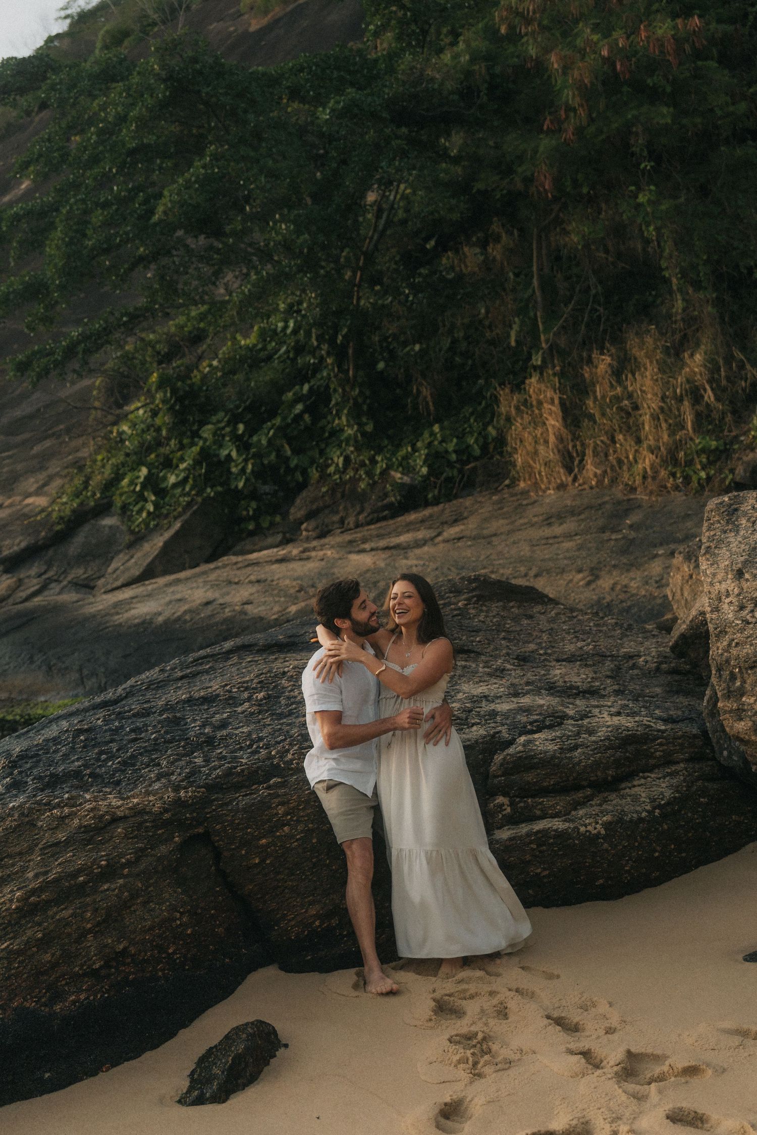Two people share an intimate moment on a secluded beach near dramatic rock formations at sunset.
