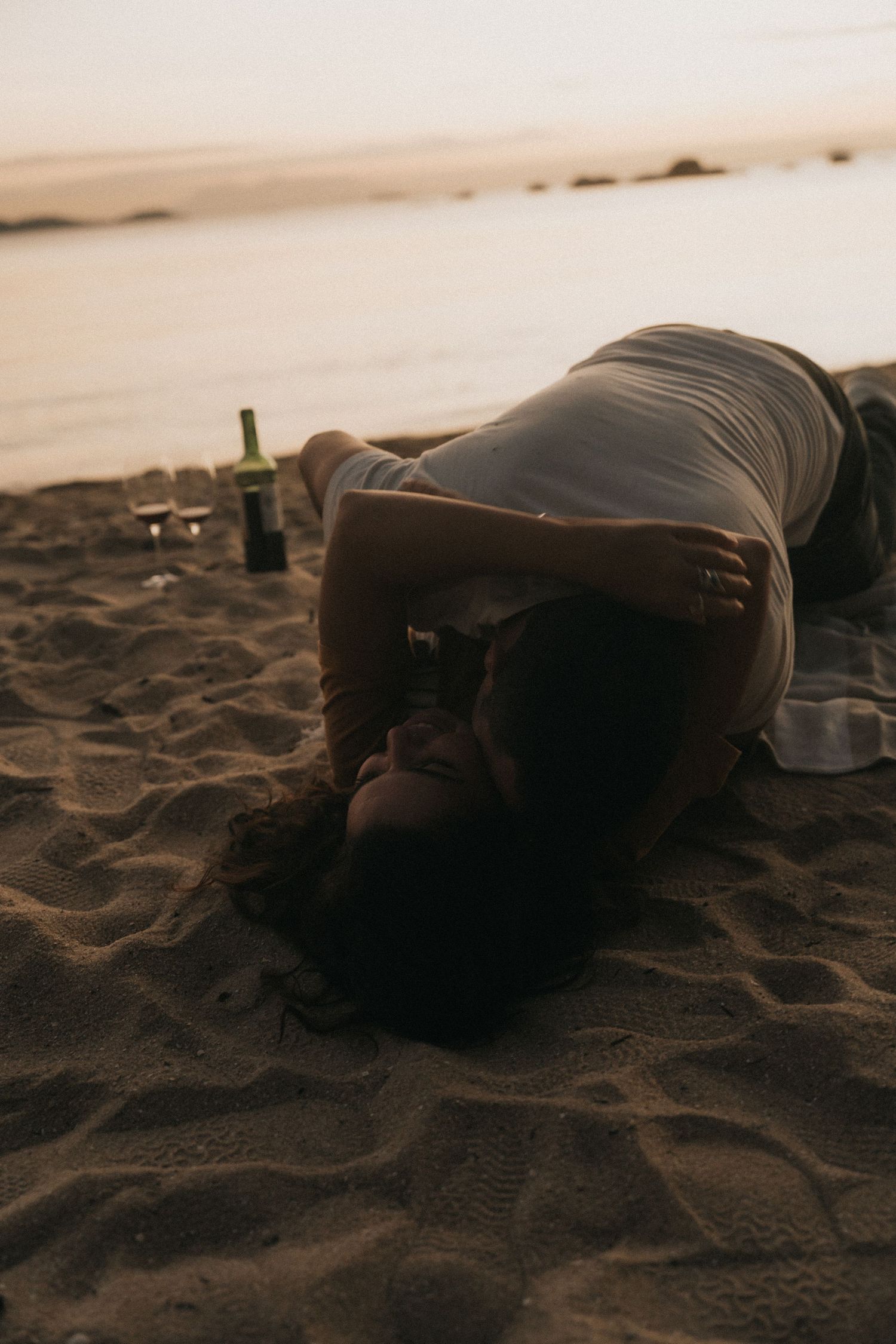 A person lies still in the dark on a sandy beach, shown in a sequence of five similar photos at sunset.