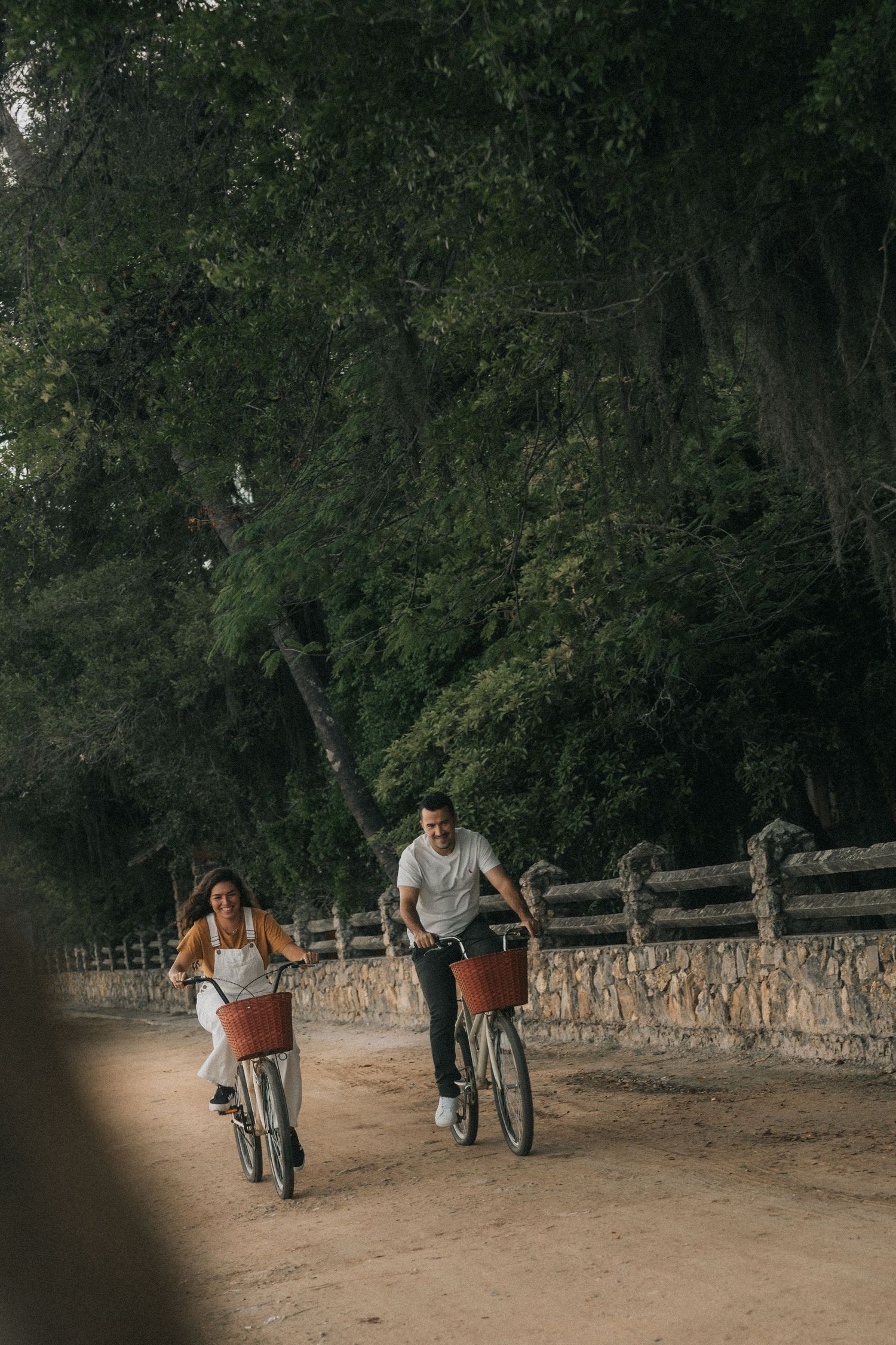 A couple riding bicycles on a dirt path along a rustic wooden fence in a wooded area during sunset.