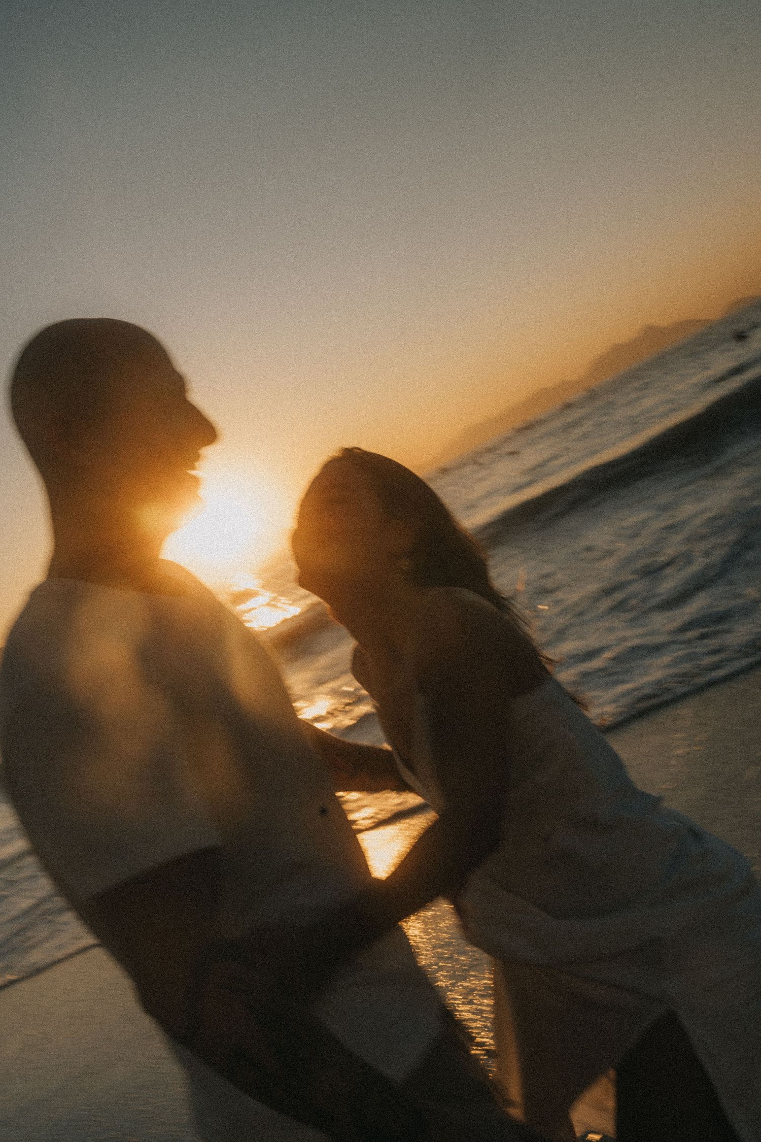A silhouetted couple sits together on a beach during sunset, creating a romantic atmosphere against the ocean backdrop.