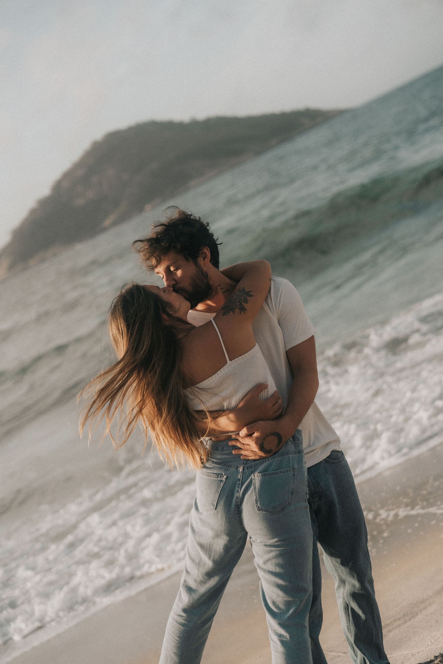A romantic couple embraces on a misty beach with crashing waves and cliffs in the background.