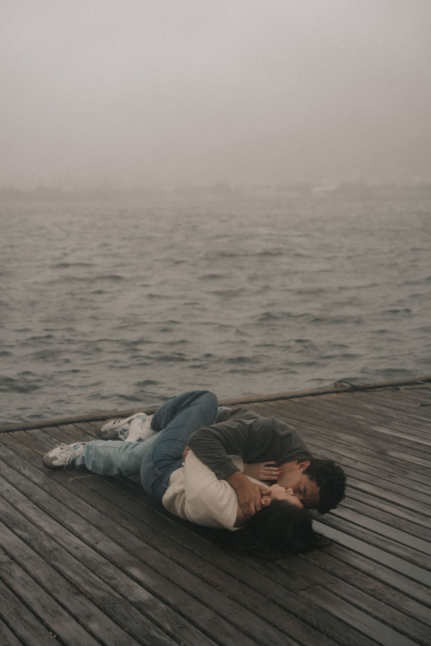 A figure lies still on a wooden dock overlooking a misty body of water as fog rolls across the scene.