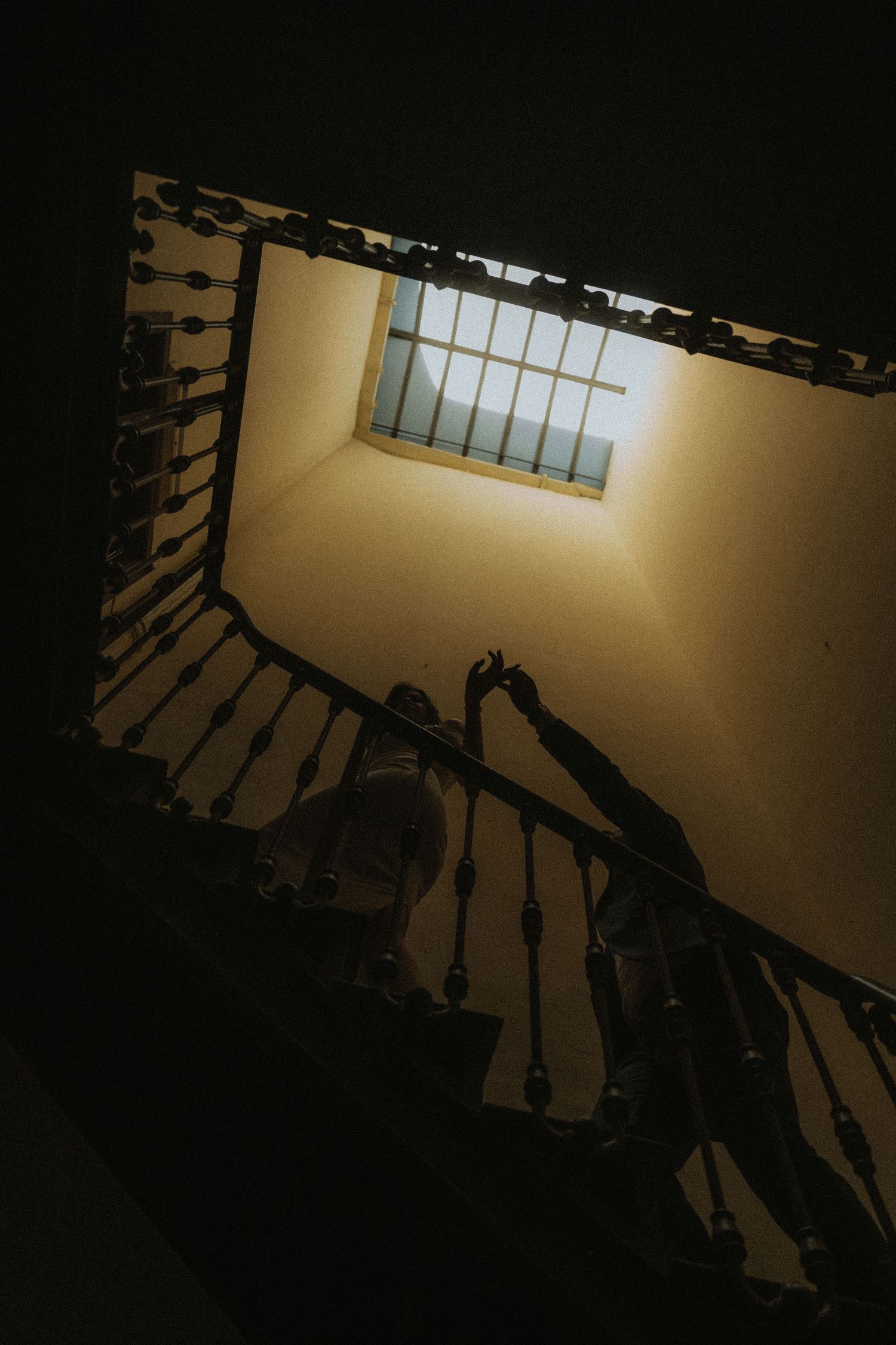 Looking up a spiral staircase with dark wooden railings against beige walls illuminated by natural light from above.