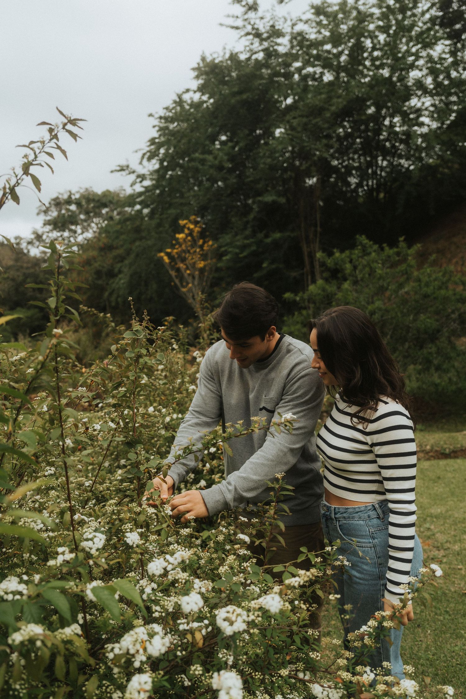 A couple examines white flowers growing in a garden during an outdoor photoshoot.