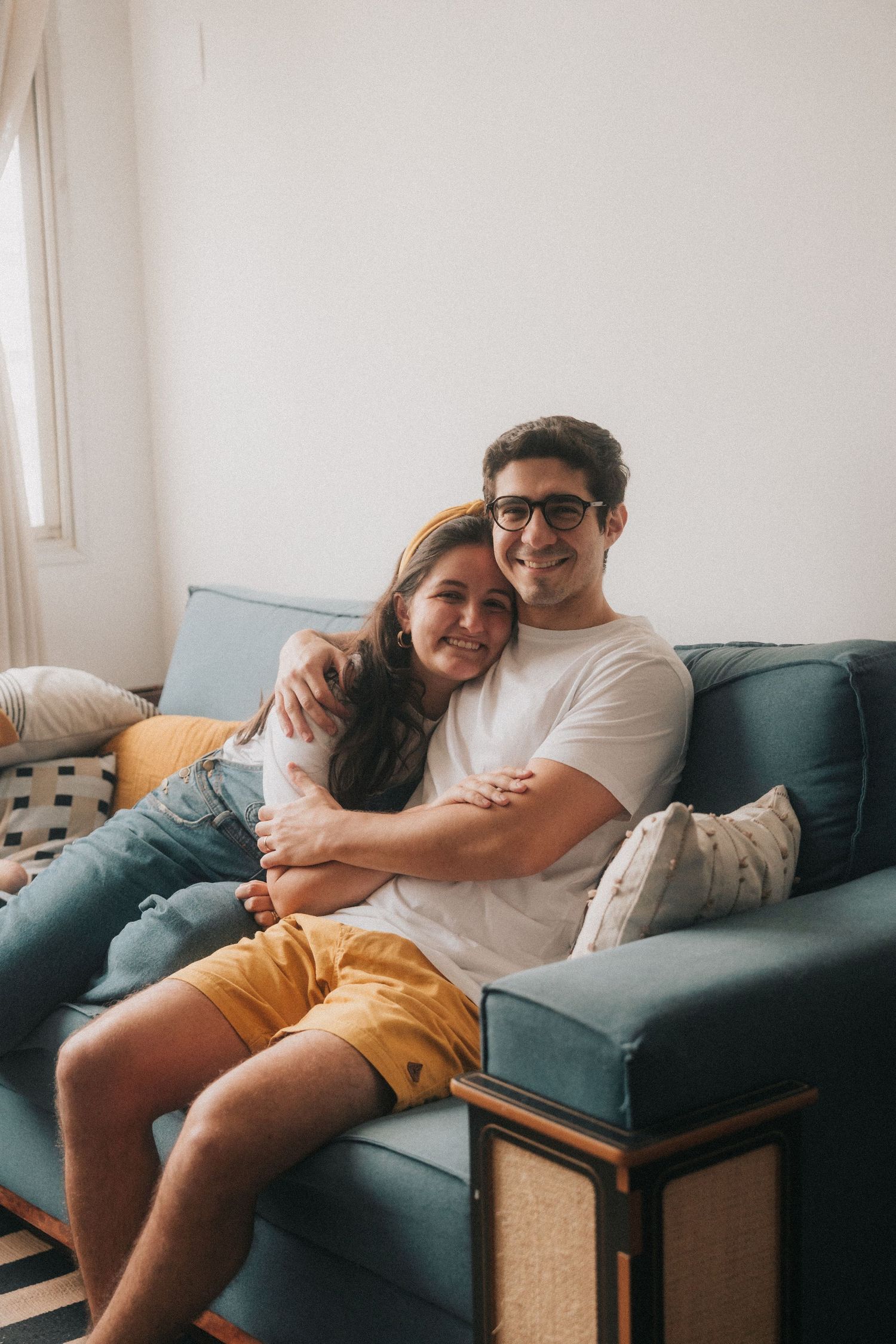 Two people cuddle together intimately on a teal blue couch in a bright sunlit room.