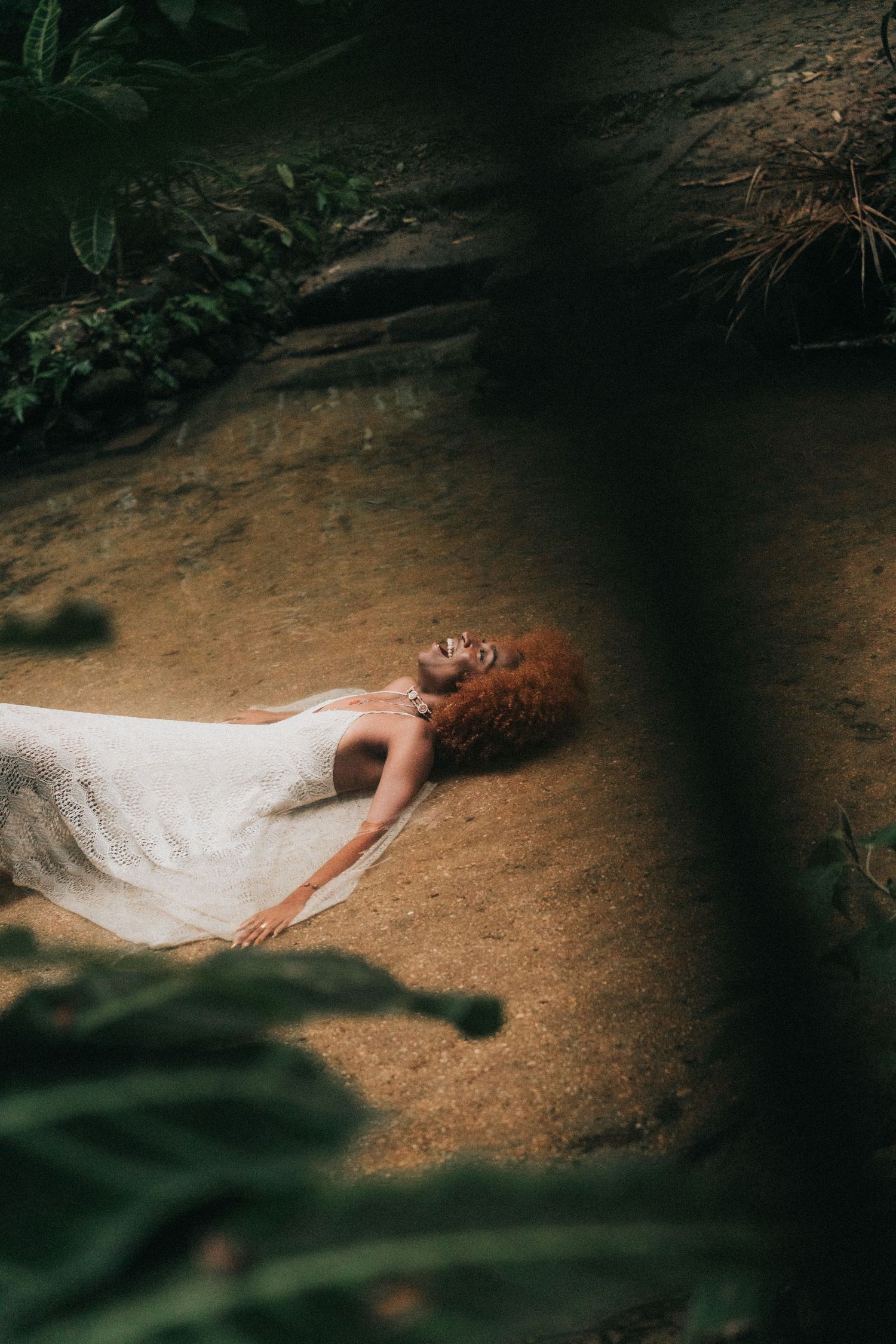 A person in a white dress lies peacefully on brown earth surrounded by dark tree shadows and foliage.