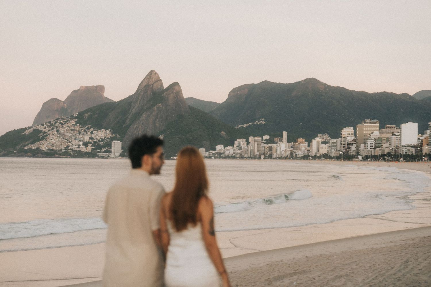 Couple walking along Copacabana beach with mountains and cityscape visible in the background at sunset.