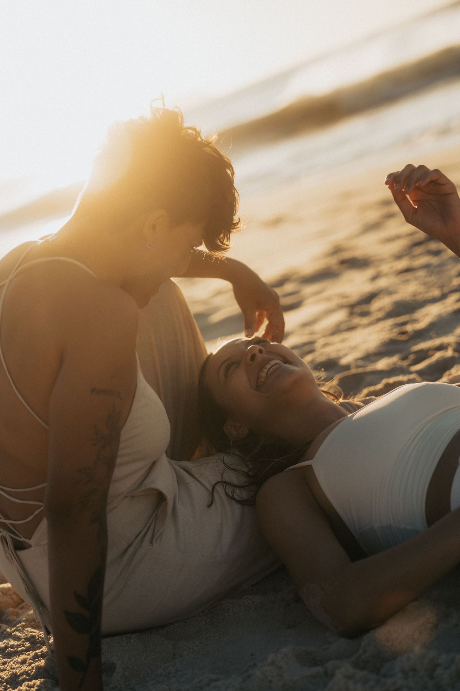 Two people share an intimate moment while lying on a sandy beach at sunset with sun flare creating a warm golden glow.