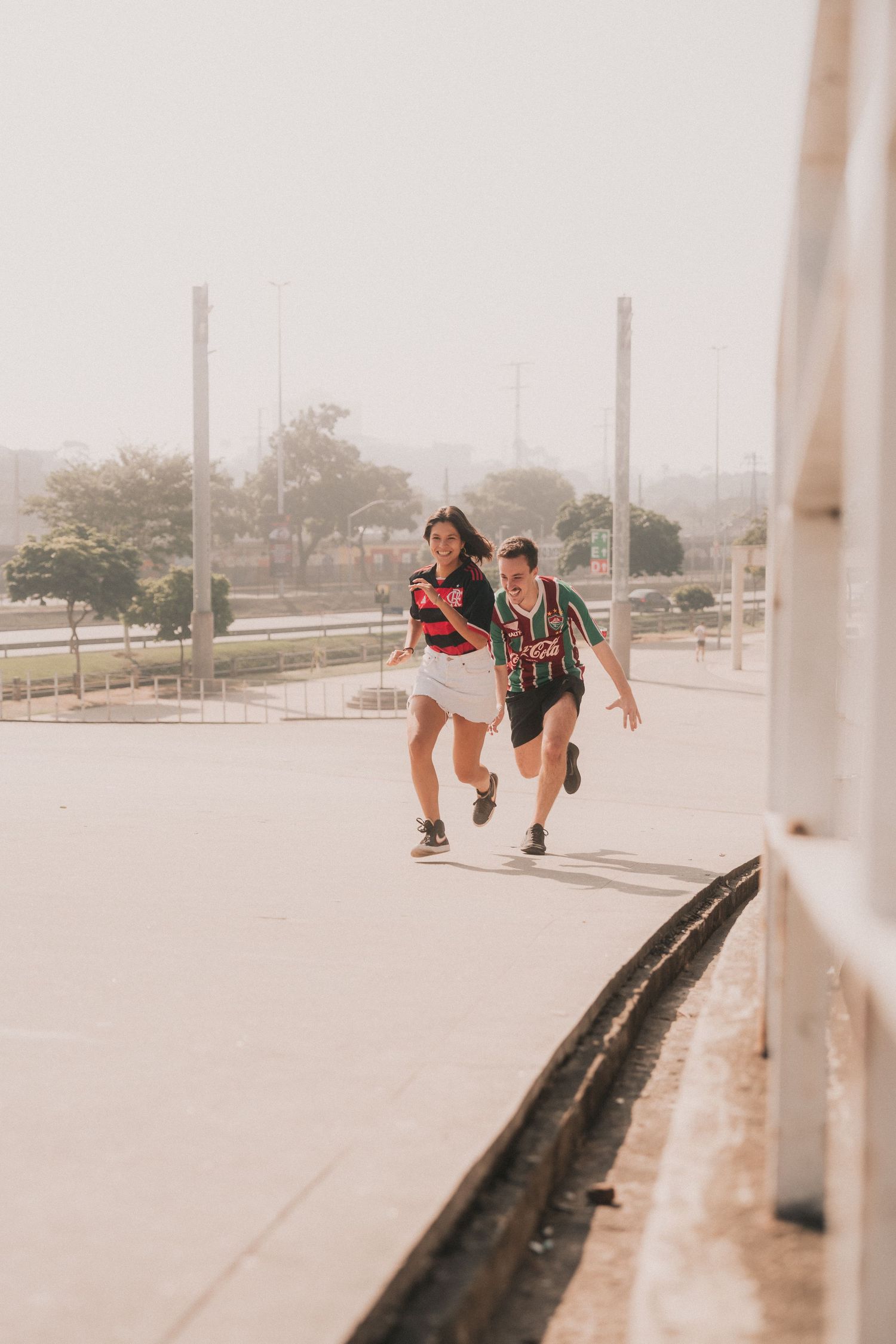 Two skateboarders joyfully riding together on a bright concrete path with urban infrastructure in the background.