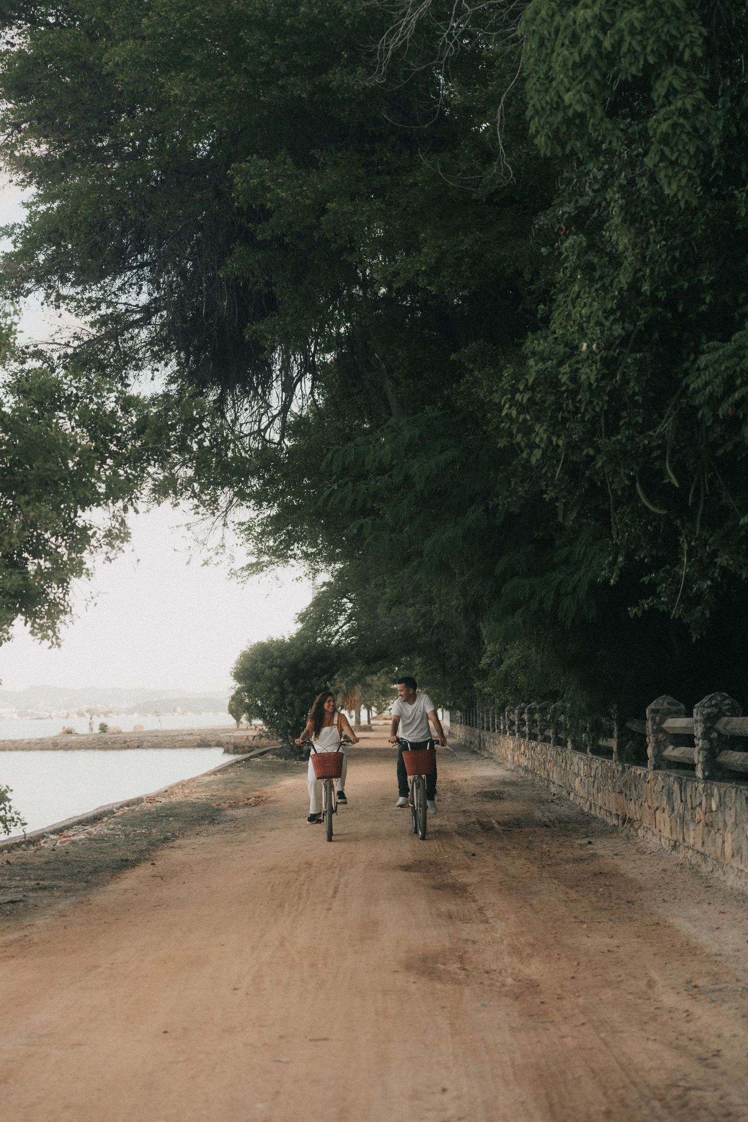 People walking and cycling on a misty dirt path lined with trees and wooden fencing on a calm morning.