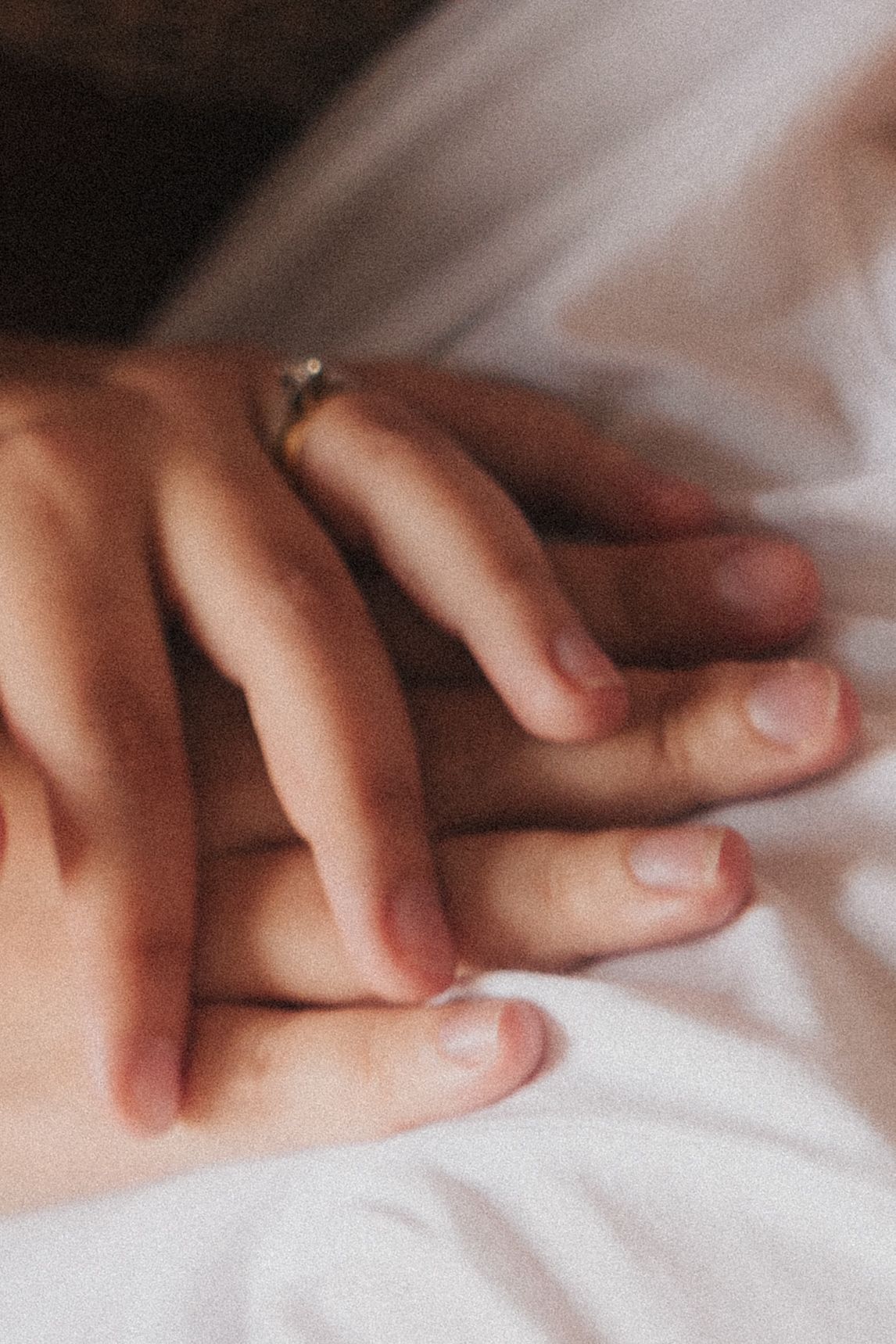 Close-up macro image of intertwined hands resting on soft white fabric with gentle natural lighting.