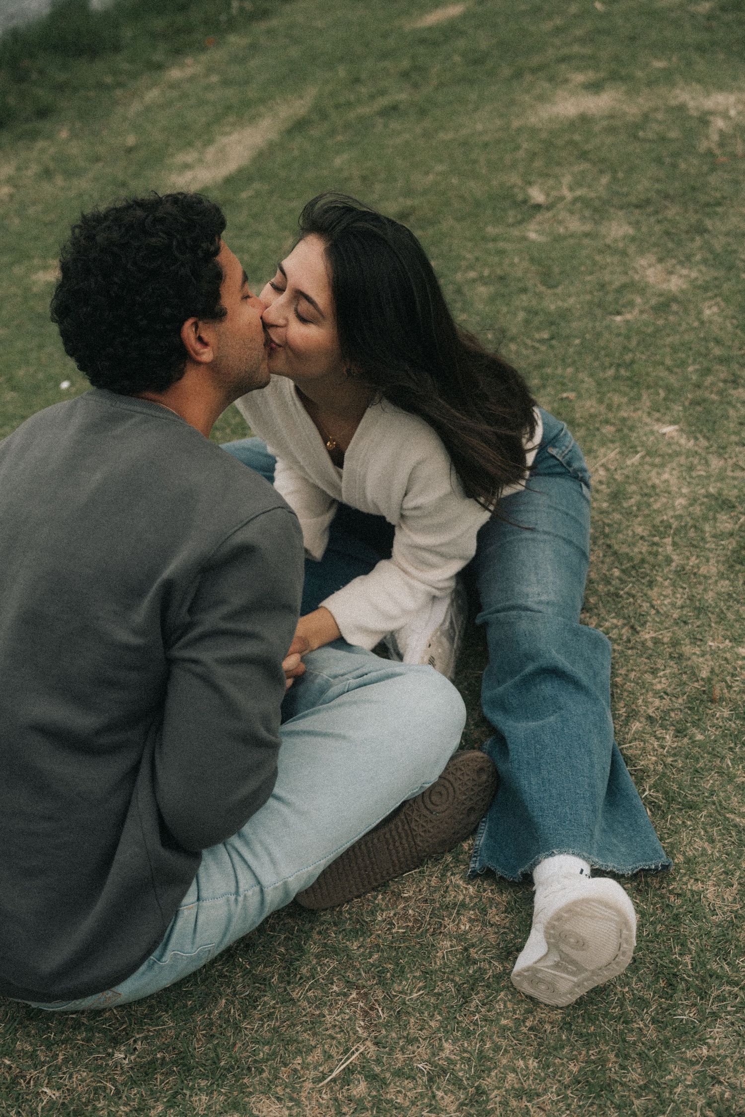 Two people share an intimate moment laying on grass outdoors while wearing casual clothing.