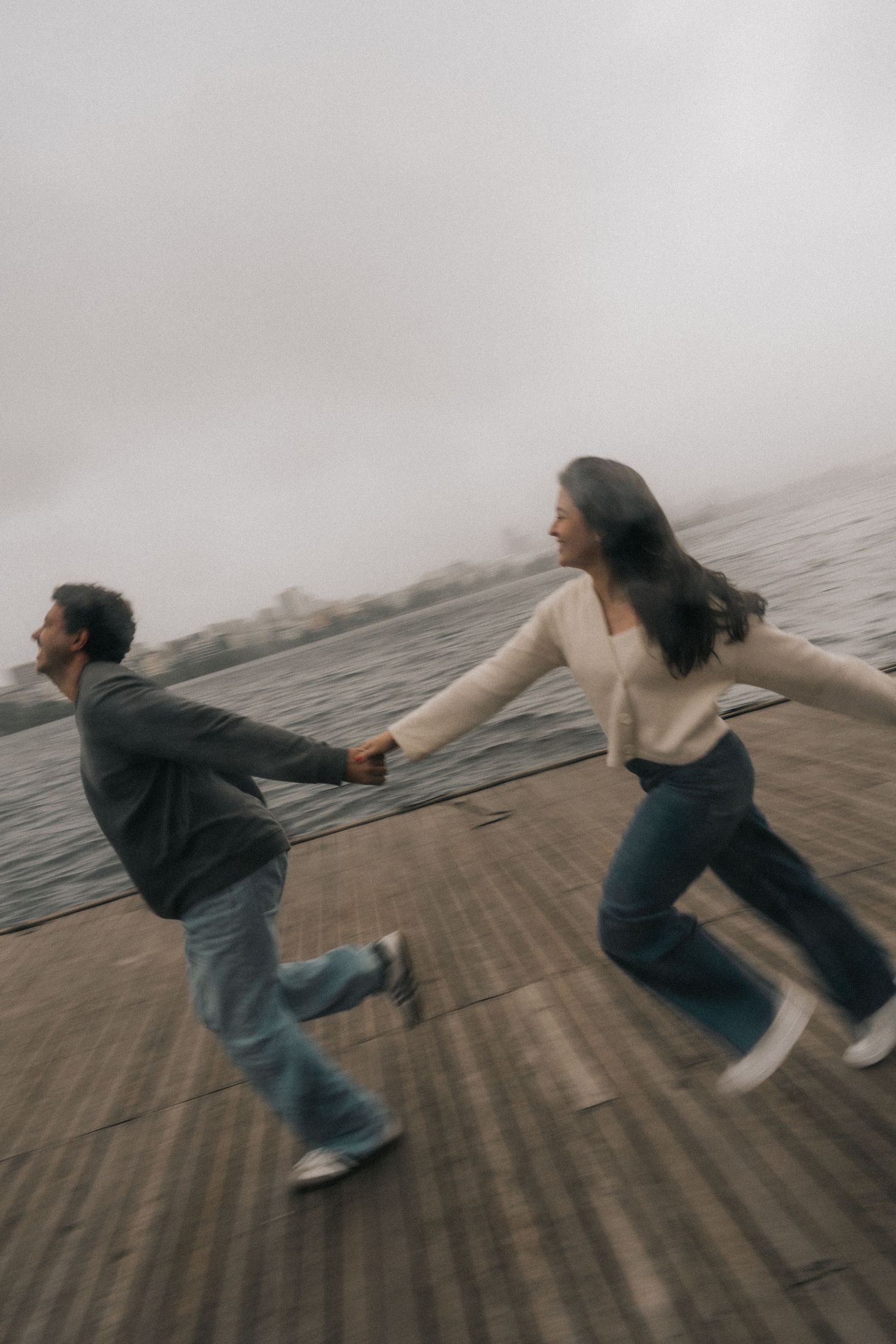 Two dancers perform a synchronized falling motion on a wooden dock by the water in moody weather.