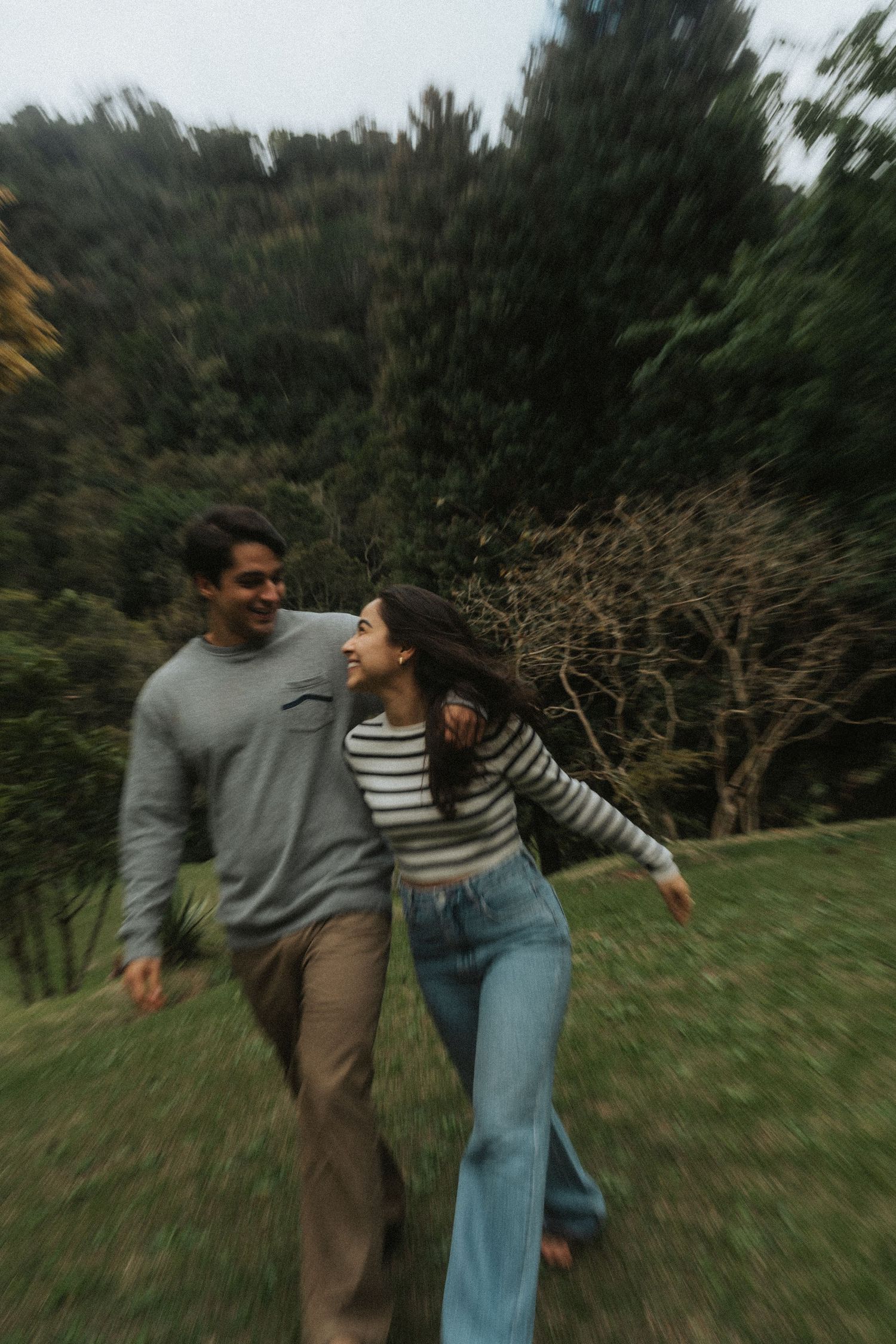 A couple in casual attire spins and dances together playfully in a grassy outdoor area with trees in the background.